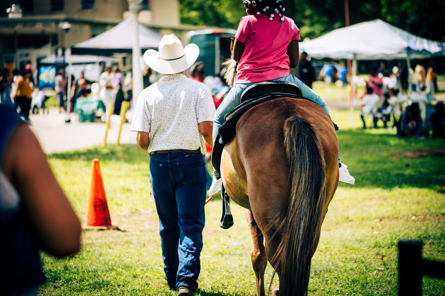 A child rides a brown horse led by a cowboy at an outdoor equestrian event on a sunny day.