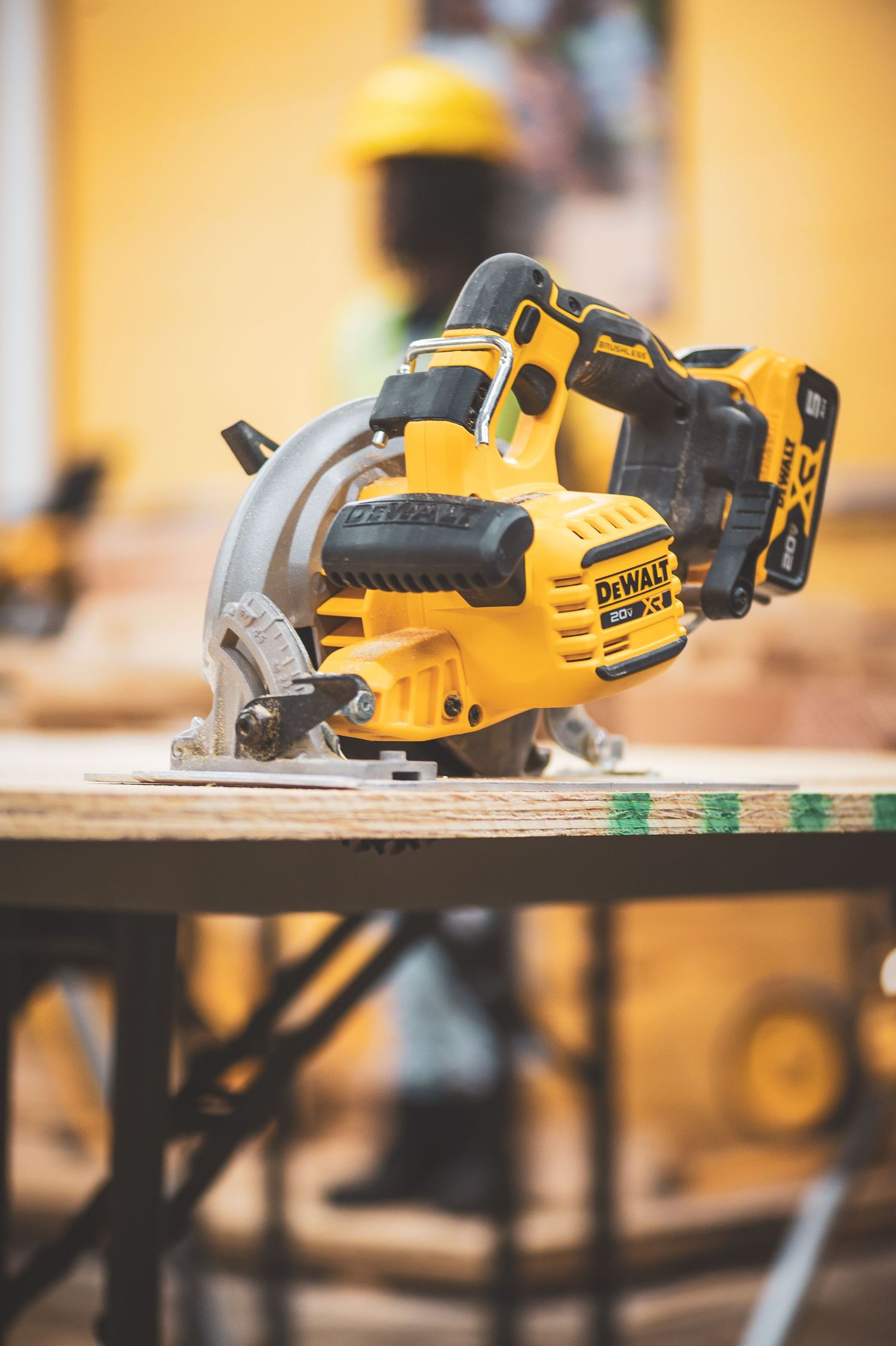 Yellow DeWalt cordless circular saw on lumber at a construction site with a worker in background.
