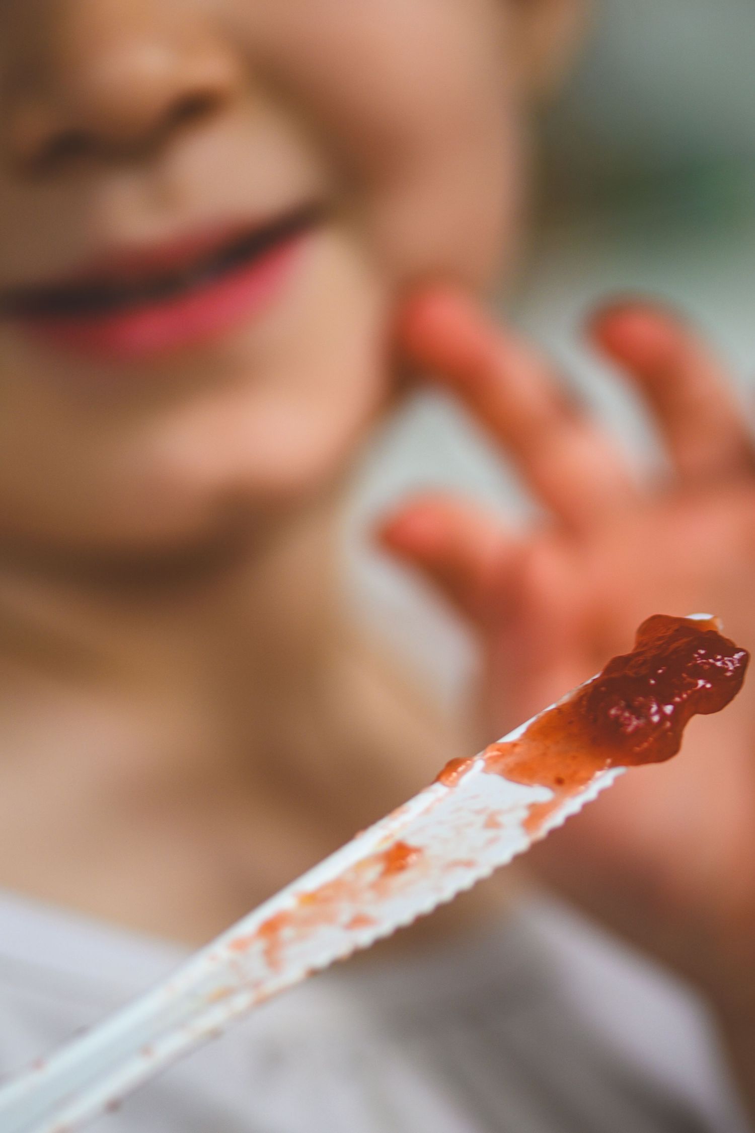 Close-up of a child holding a chopstick with red sauce, smiling in the background with messy hands.