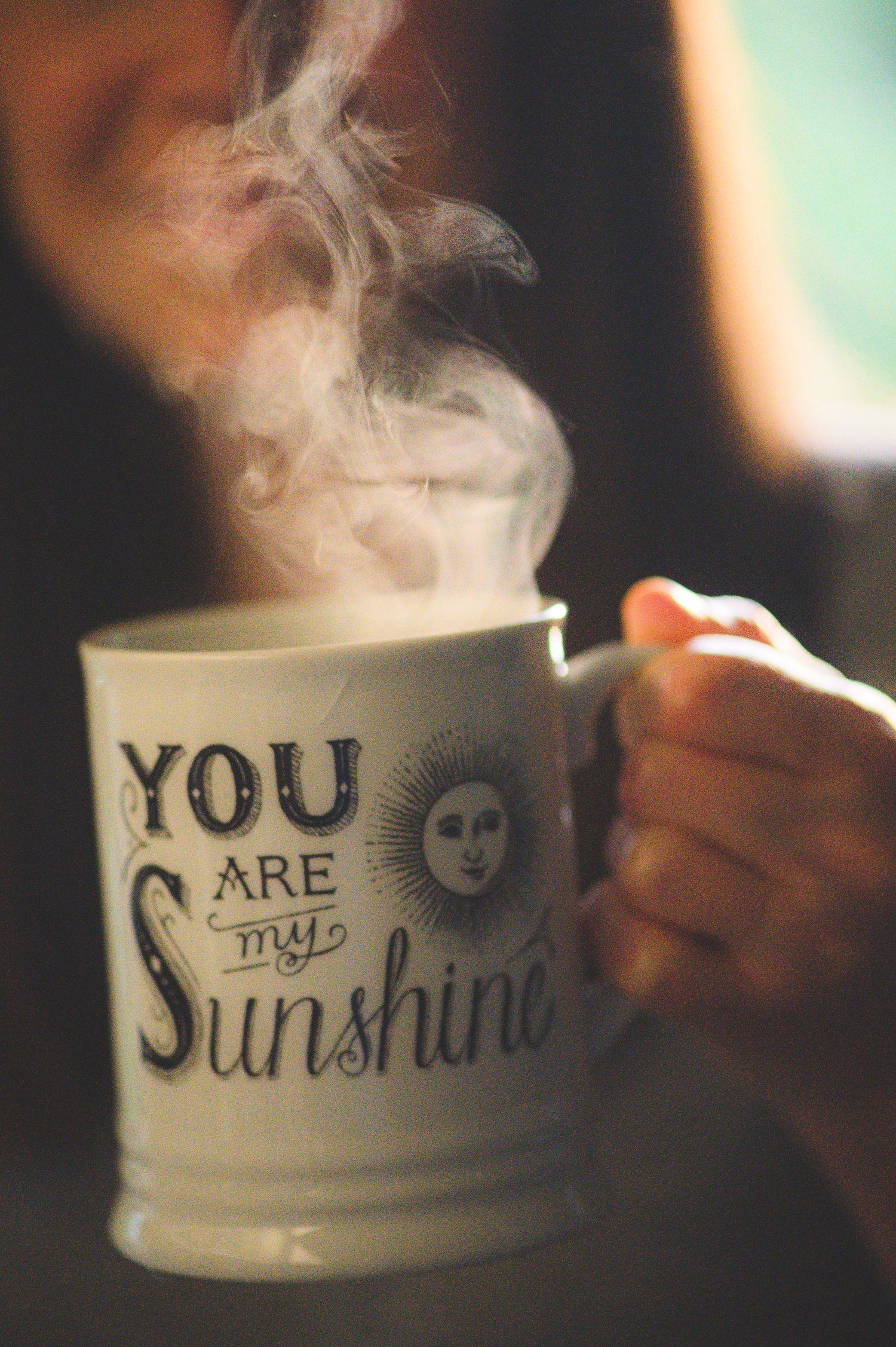 A steaming white mug with 'You Are My Sunshine' text and sun illustration held by a person.