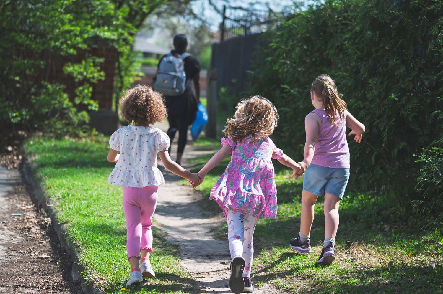 Three young girls holding hands walking down a sunny garden path with an adult ahead.