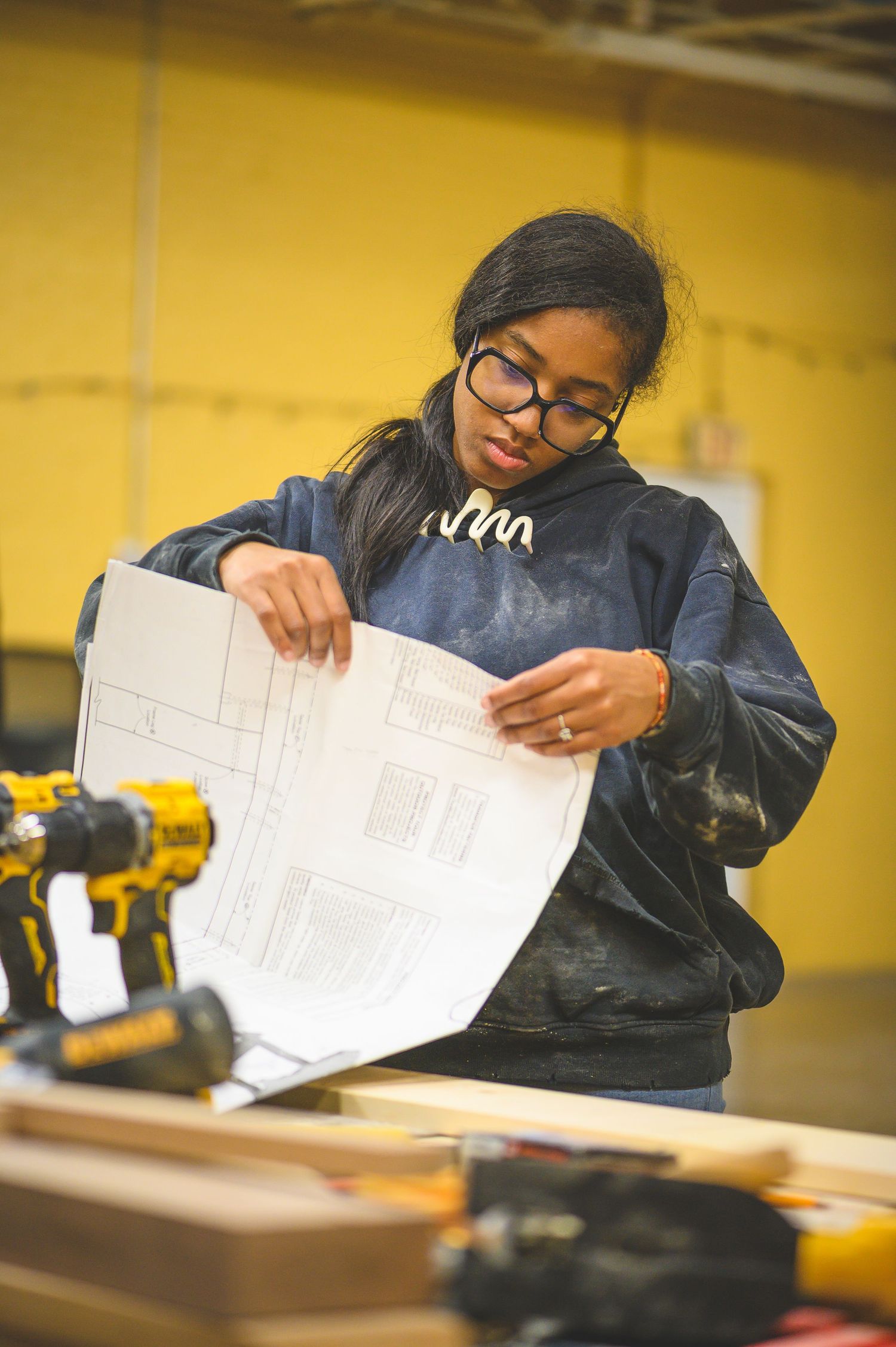 A student in a navy jacket reviews engineering blueprints at a workbench with power tools visible in the background.