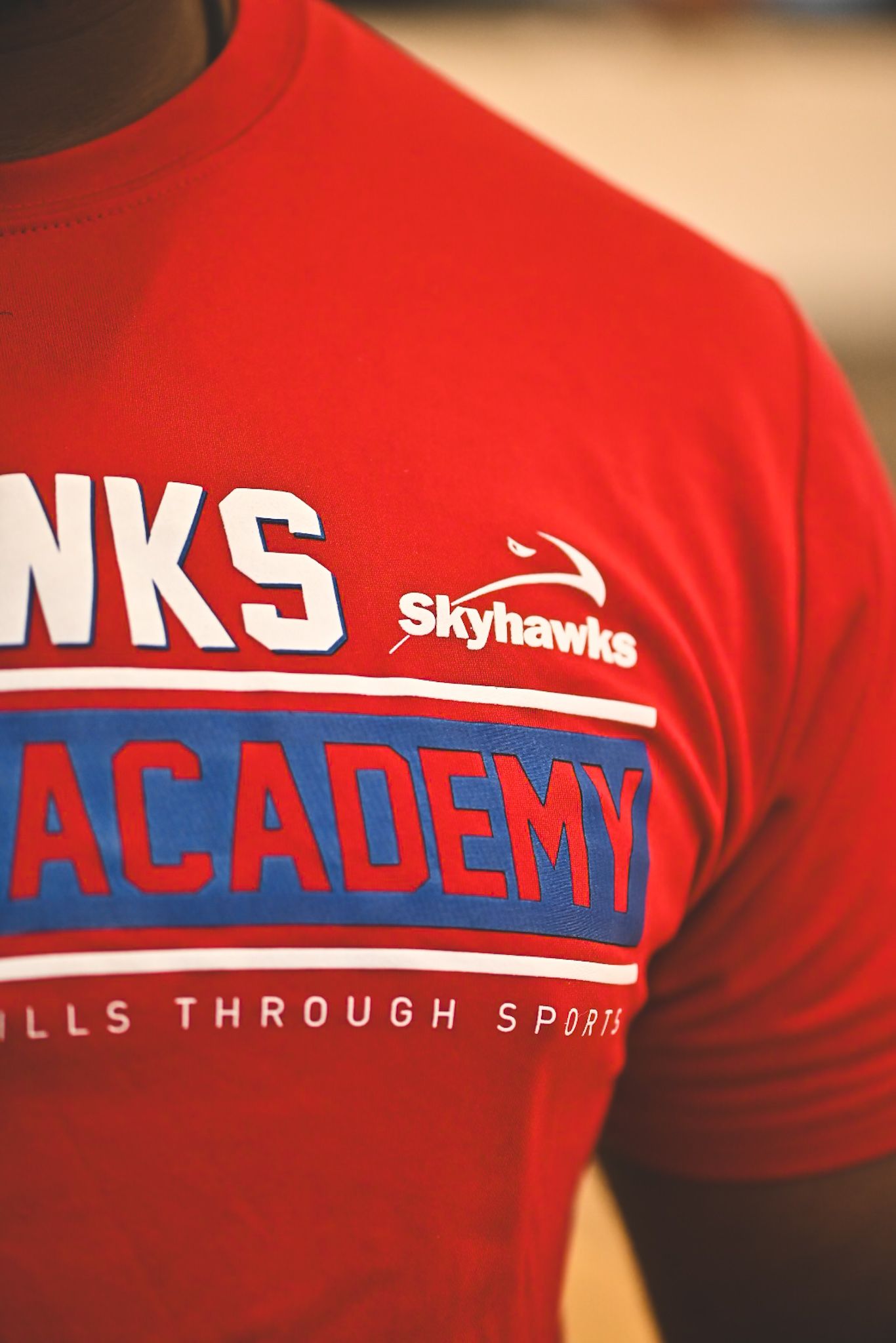 Red Skyhawks Academy t-shirt with blue and white lettering reading Hawks Academy Skills Through Sports.