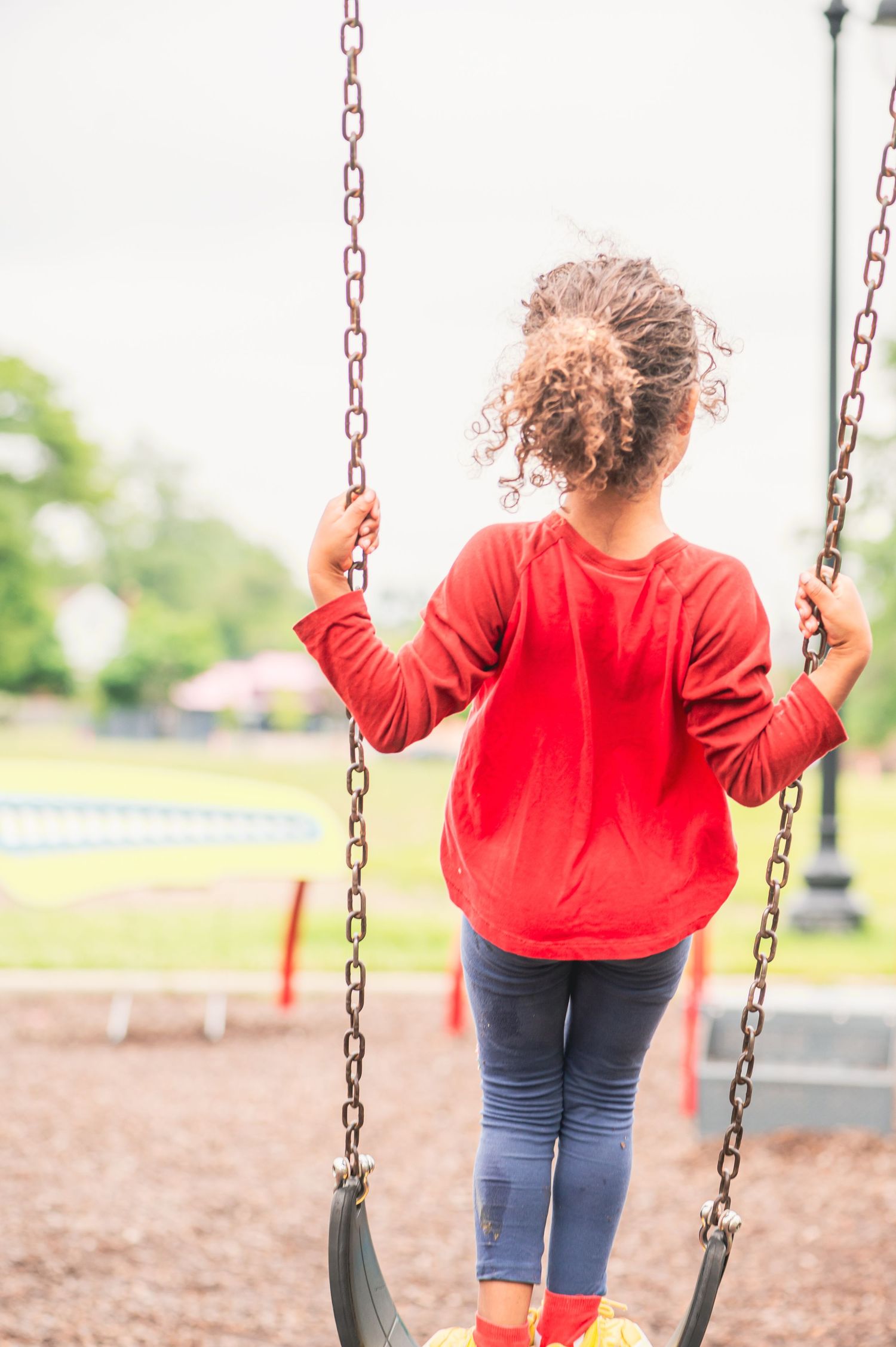 Child wearing a red sweater and jeans sits on a playground swing on a cloudy day at the park.