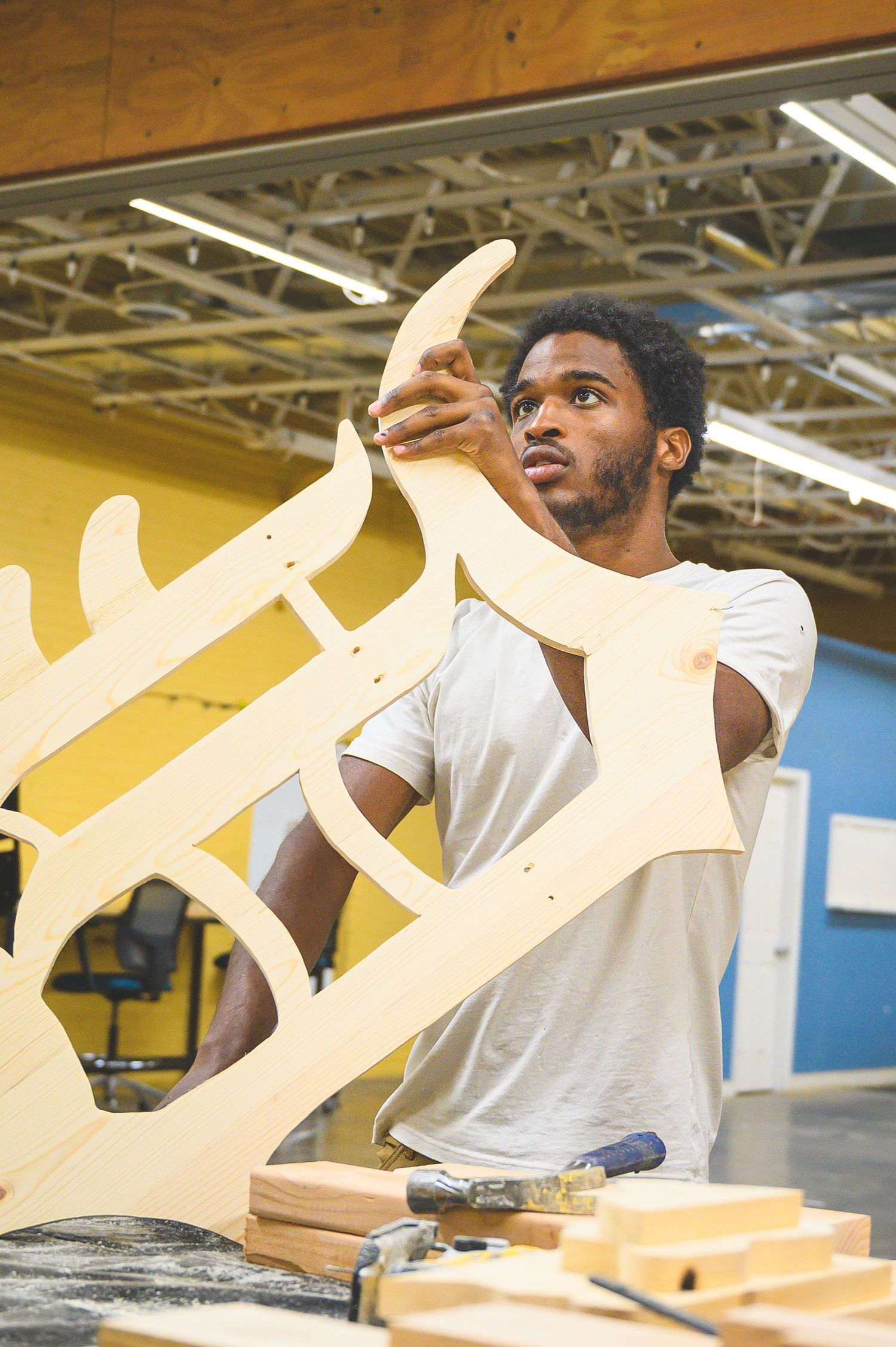 A person in a white t-shirt concentrates while working with wooden materials in a workshop with industrial lighting.