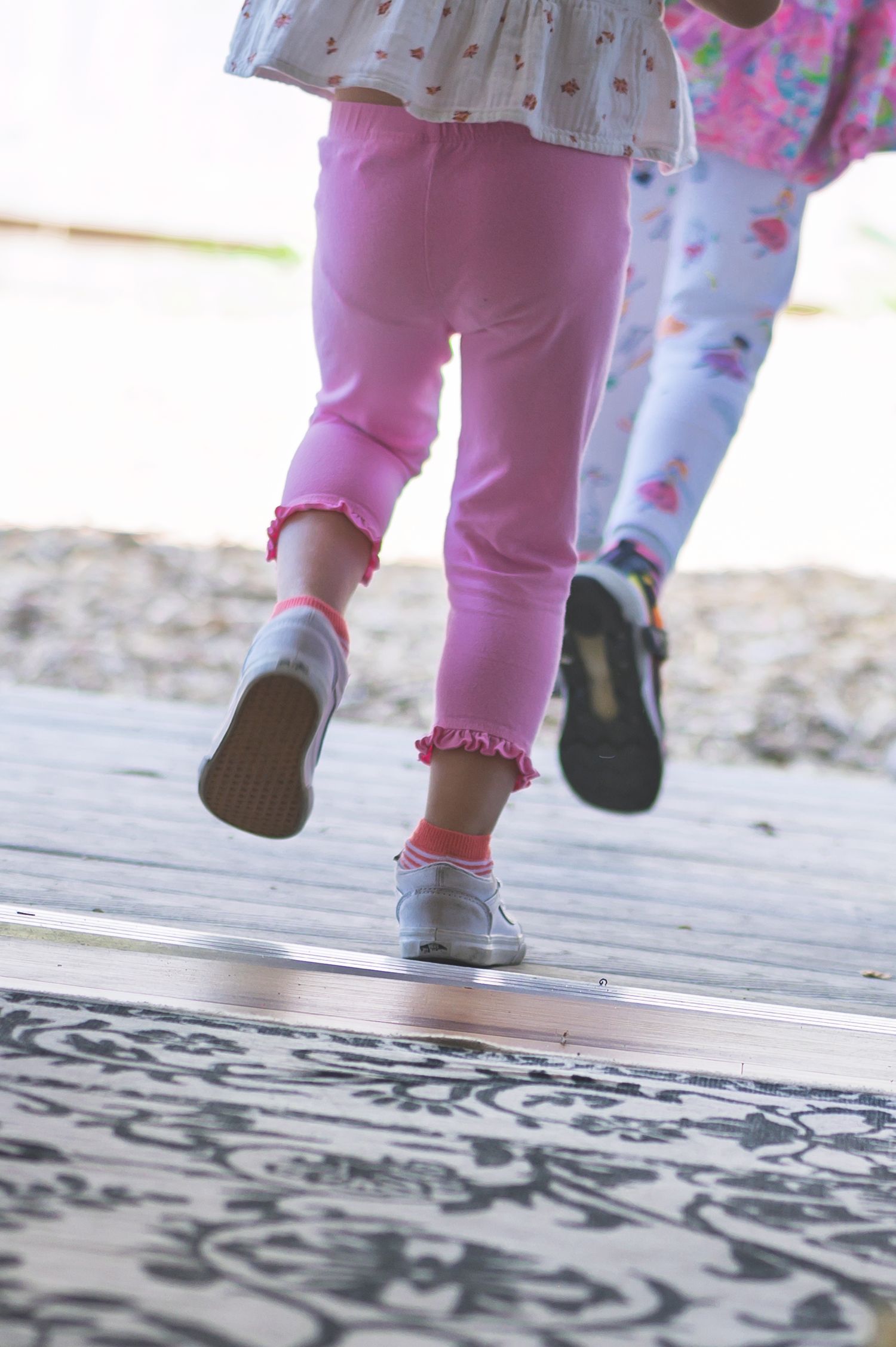 Two young girls in pink leggings and silver shoes running on a wooden deck at a playground.