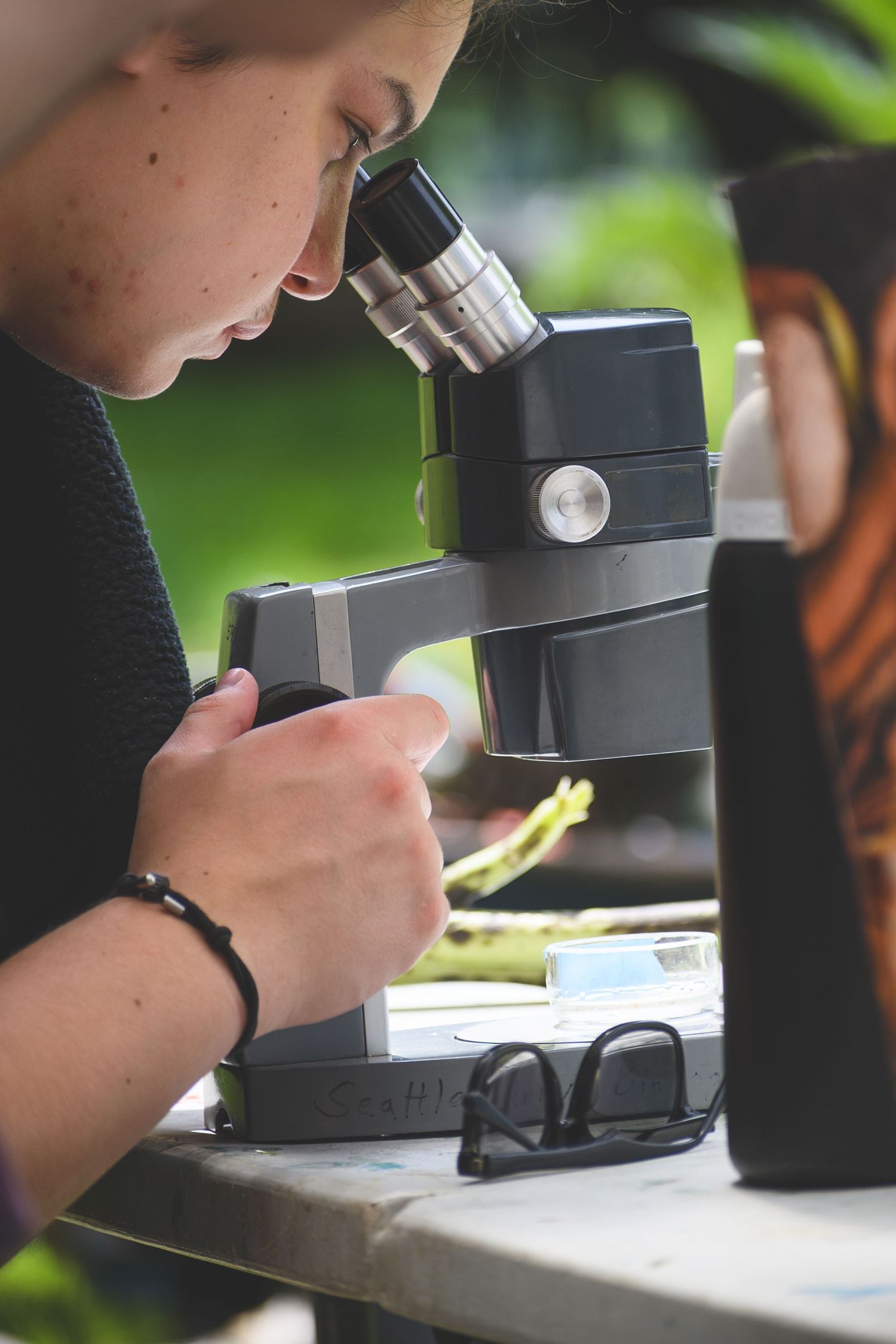 Young student peers through a microscope outdoors, conducting a science experiment at a table.
