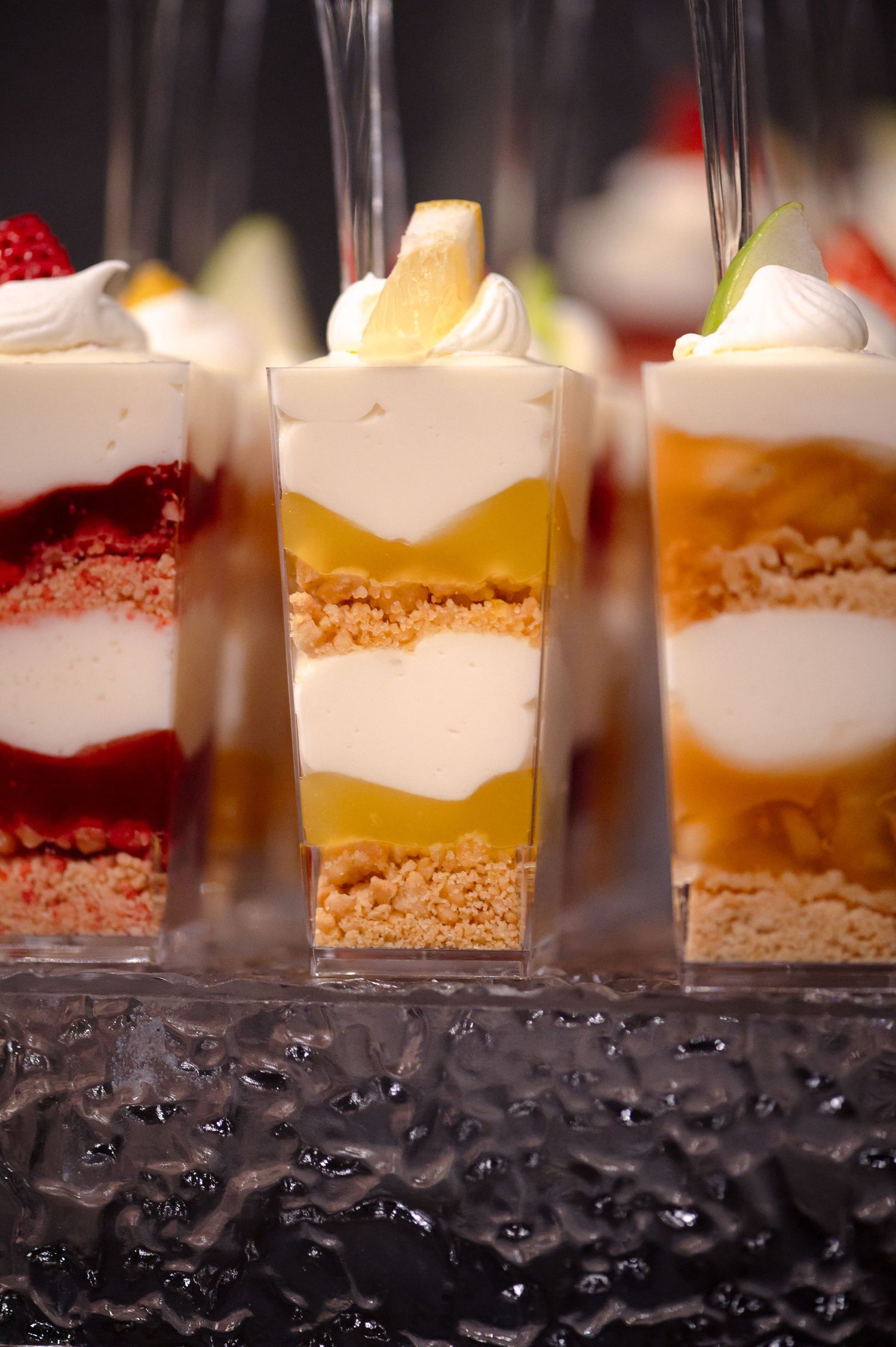 Three elegant layered dessert shooters featuring cream, fruit, and cookie crumb bases displayed in clear glasses on a textured surface.