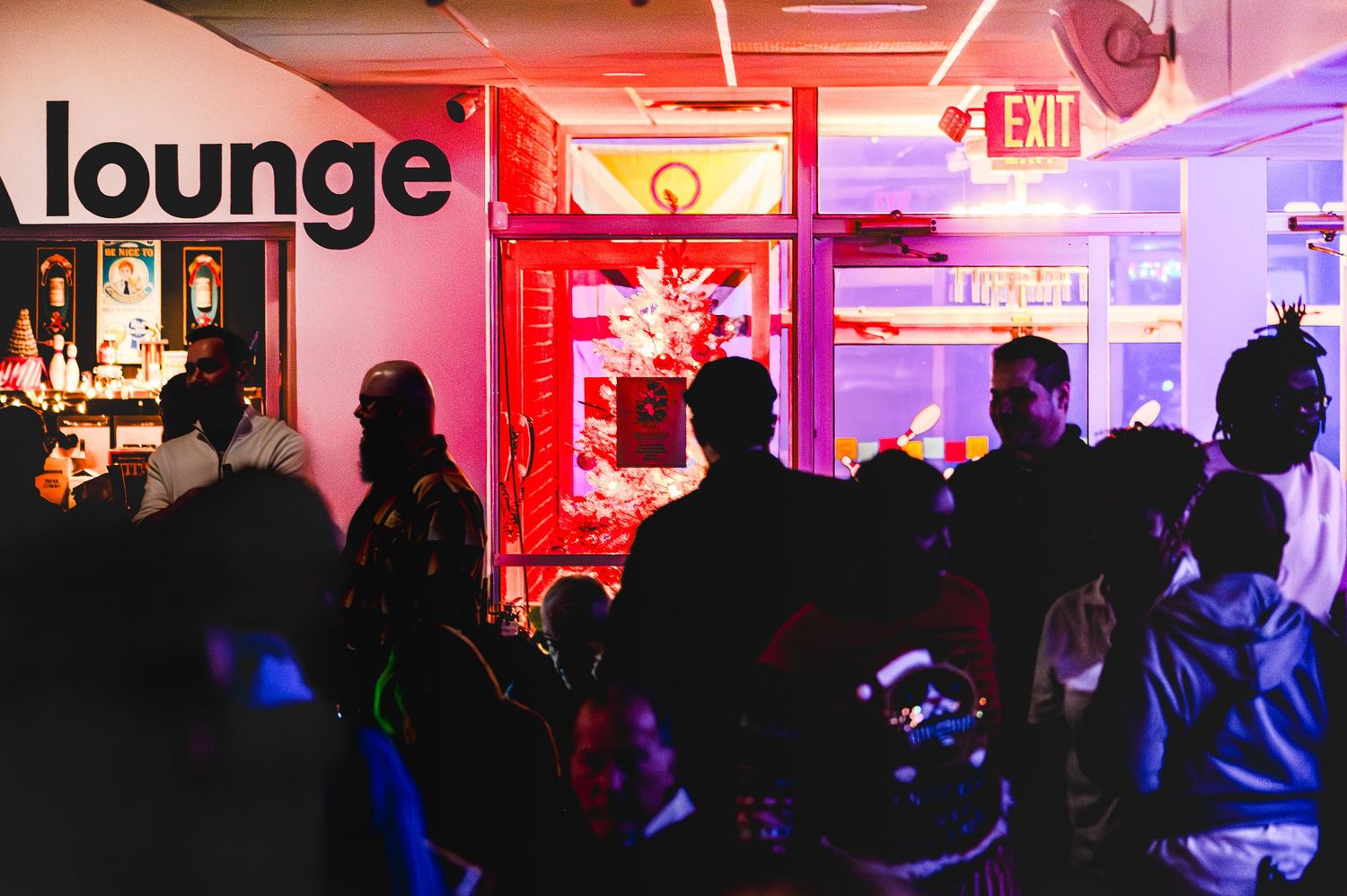 Crowded lounge bar with red neon lighting, guests socializing near entrance with exit sign glowing above.