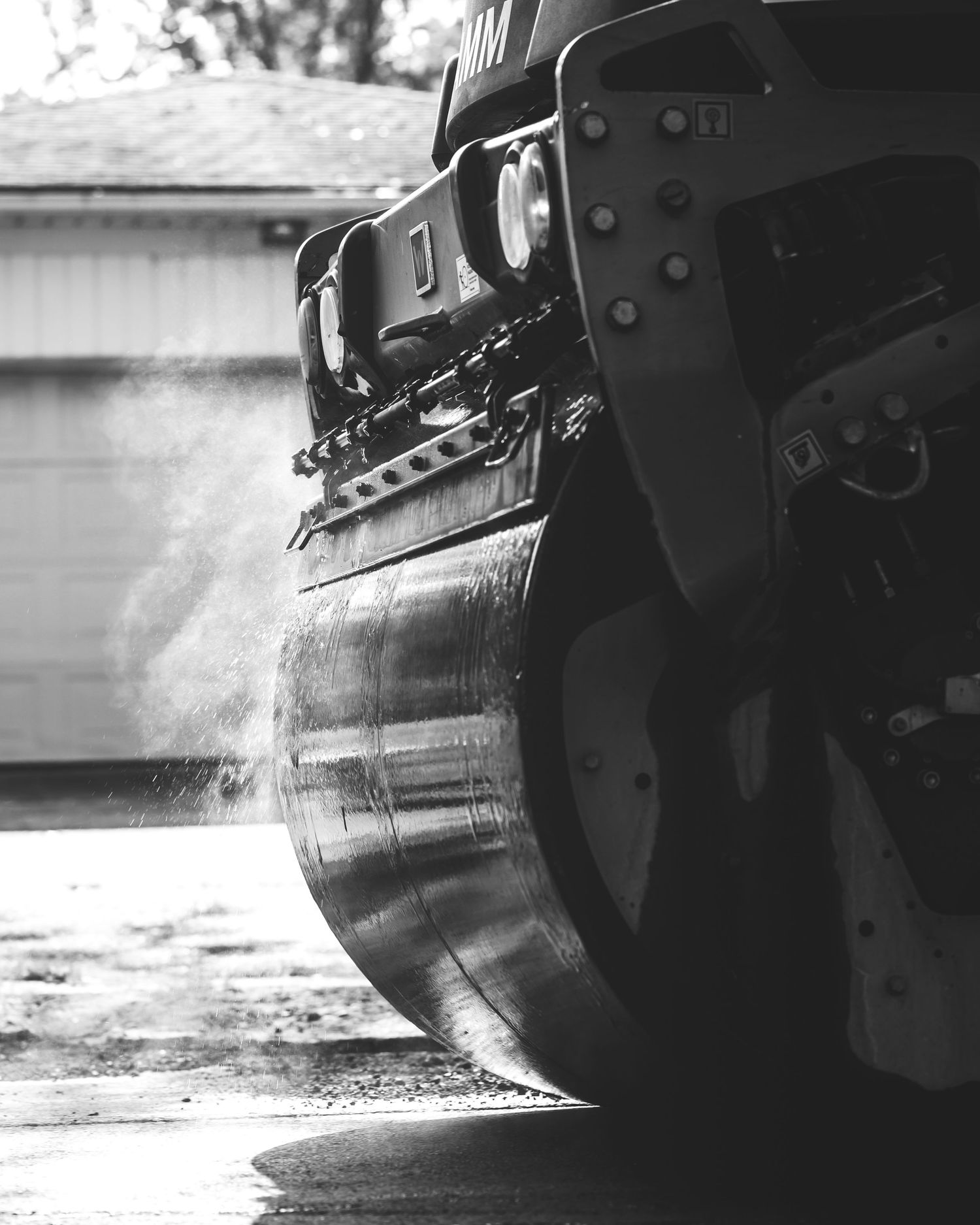 Black and white close-up of a heavy machinery roller compacting asphalt with dust rising, parked near a garage.