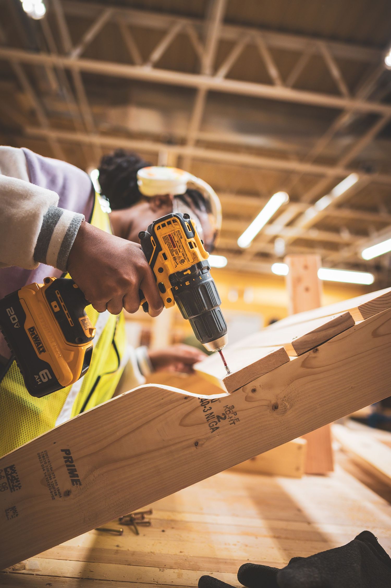 Worker in safety vest using a yellow DeWalt cordless drill on wood framing at a construction site.