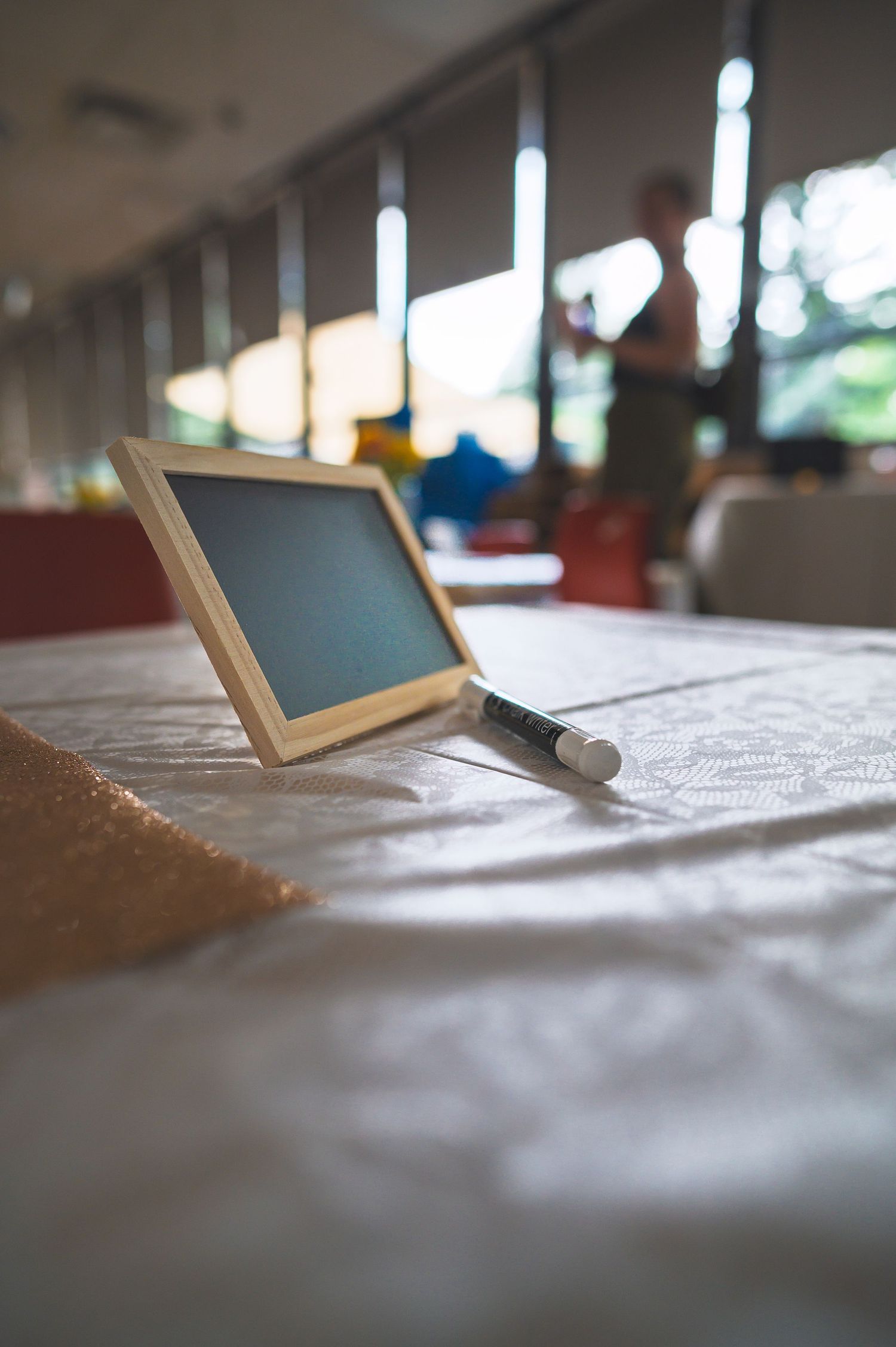 Small chalkboard with chalk on a white tablecloth in a softly lit room with blurred background figures.