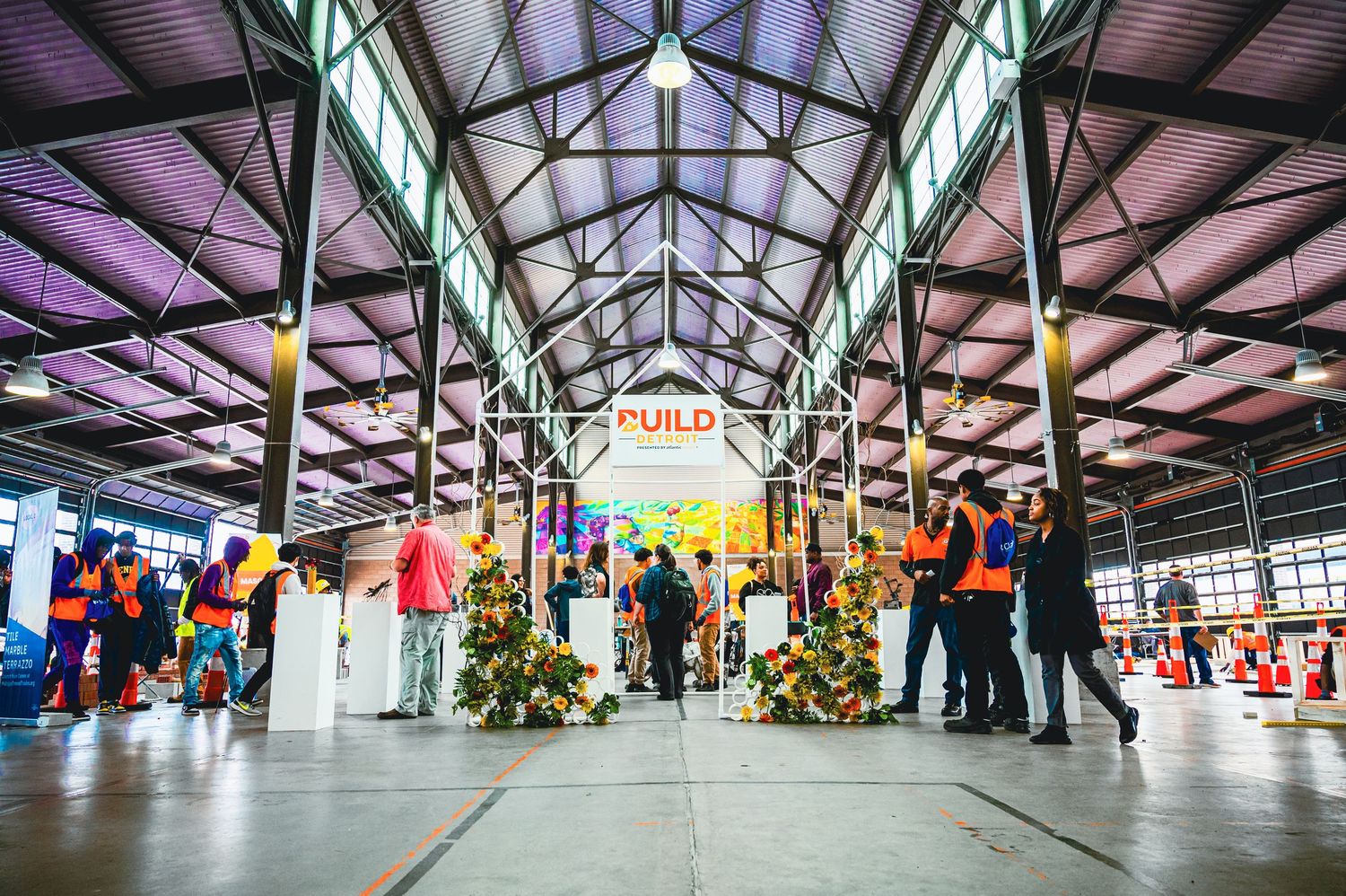 People in hi-vis vests gather inside a large industrial hall at a BUILD event with floral displays and colorful installations.