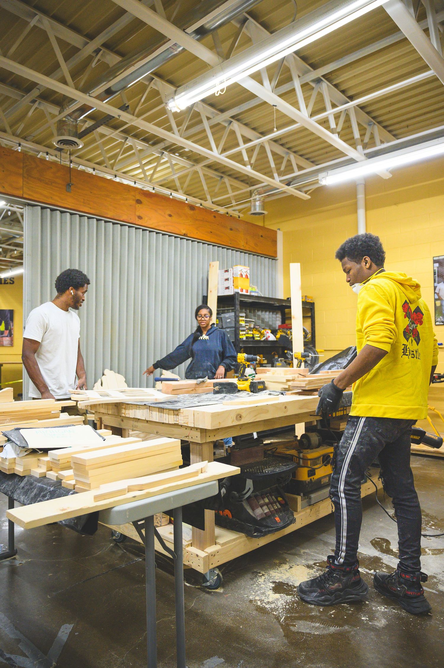 Construction workers measuring and cutting wooden beams on long work tables in a spacious industrial workshop.