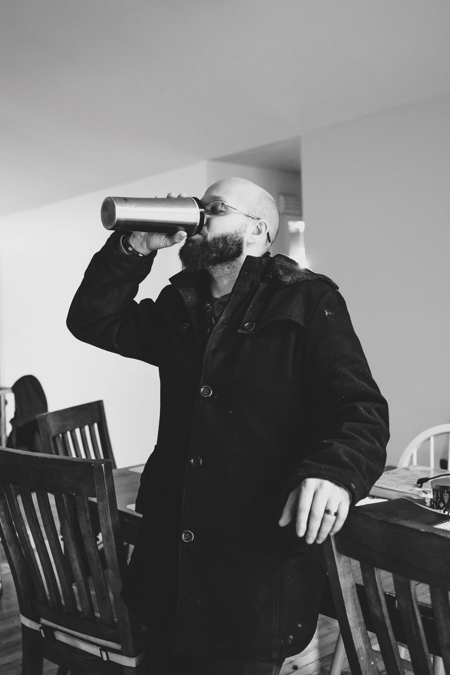 dj in a black coat drinks from a large thermos while standing indoors in this black and white photograph.