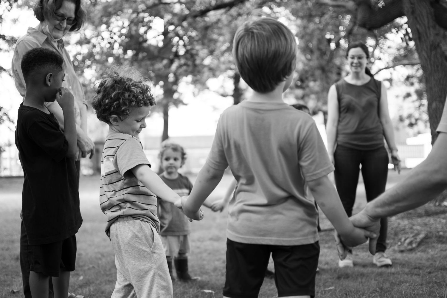 Children holding hands in a circle outdoors under trees, playing a group game supervised by an adult woman in a park.