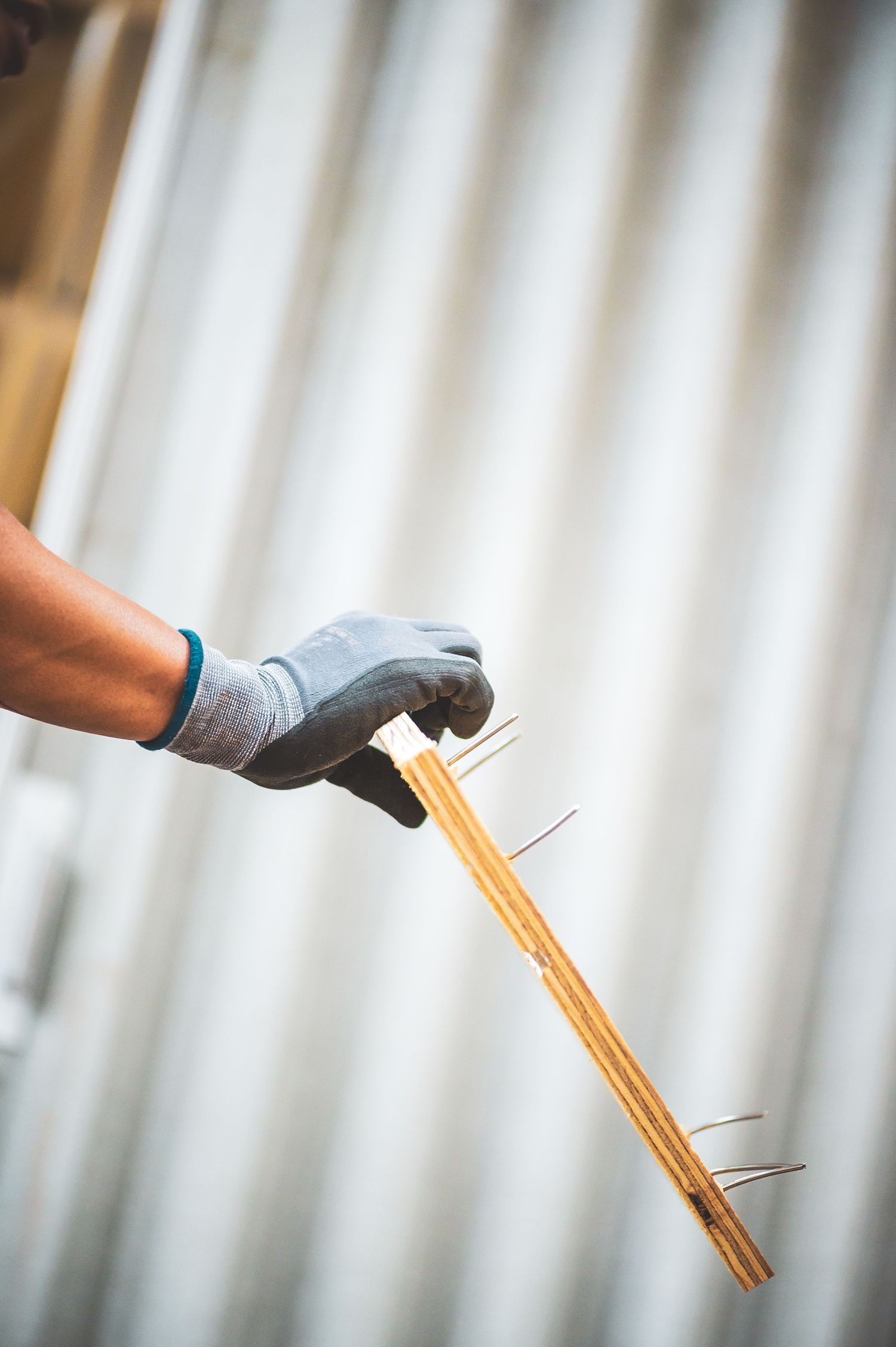 Gloved hand holding a wooden strip with protruding nails against a corrugated metal background.