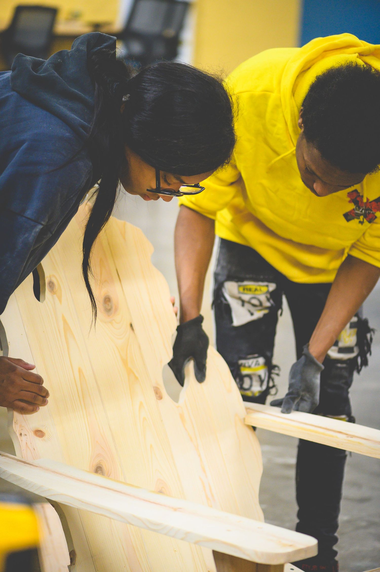Two people wearing gloves lean over to examine a piece of light-colored wood in a workshop setting with yellow walls.