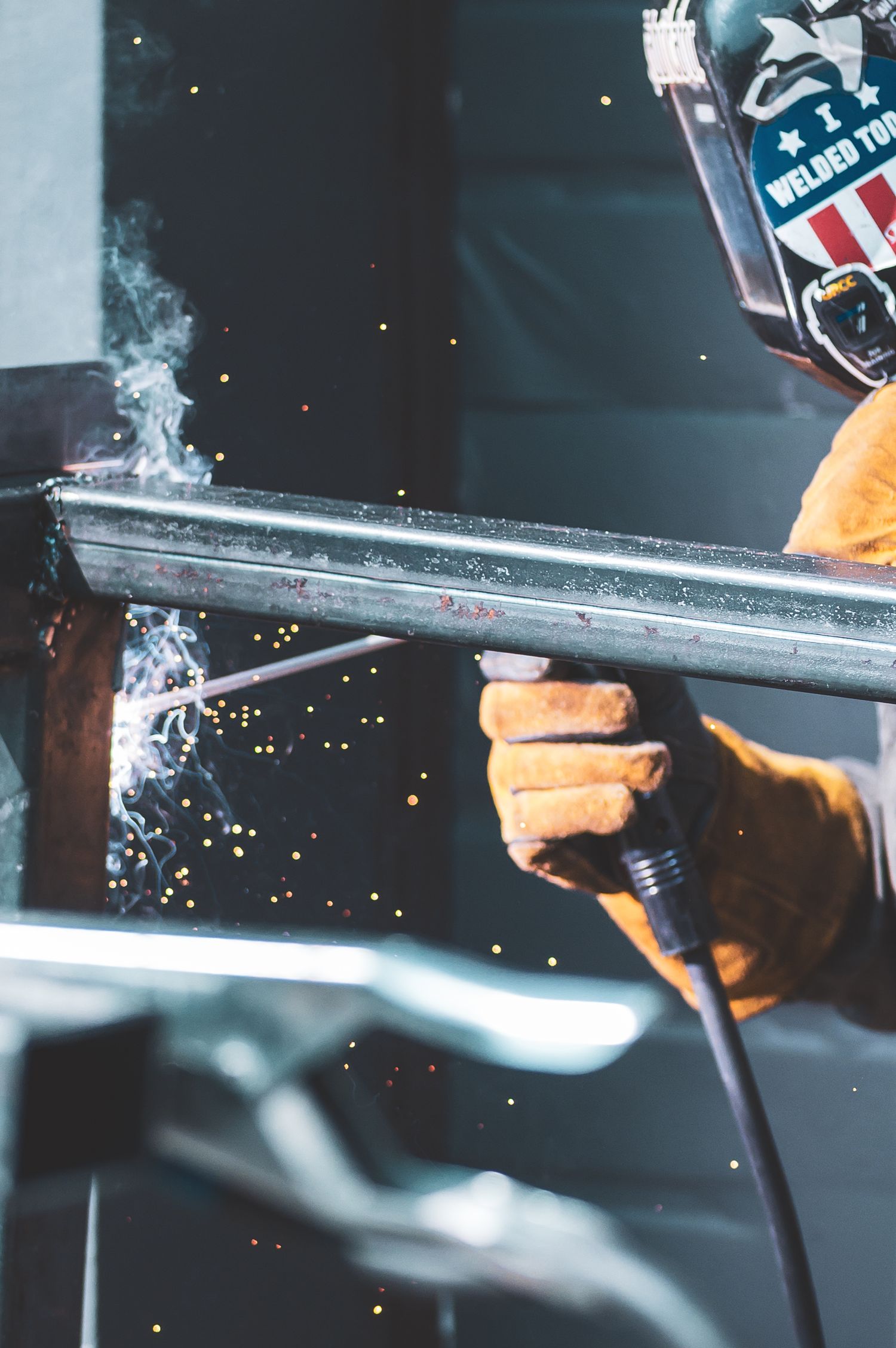 A welder in protective gear and gloves uses a welding torch on metal, with sparks and smoke flying in a workshop.