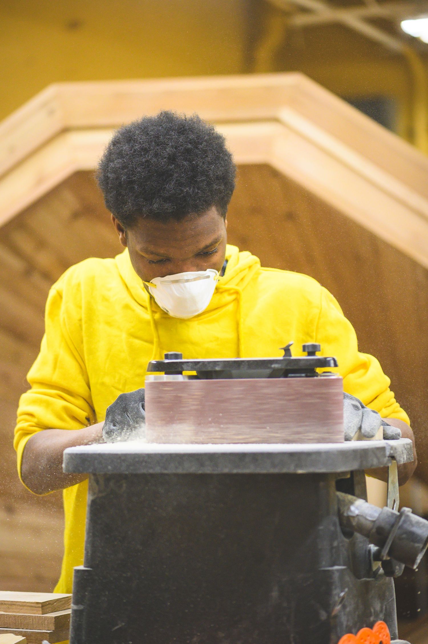 Student wearing yellow sweatshirt and protective mask works carefully on a woodworking project using power tools in workshop.
