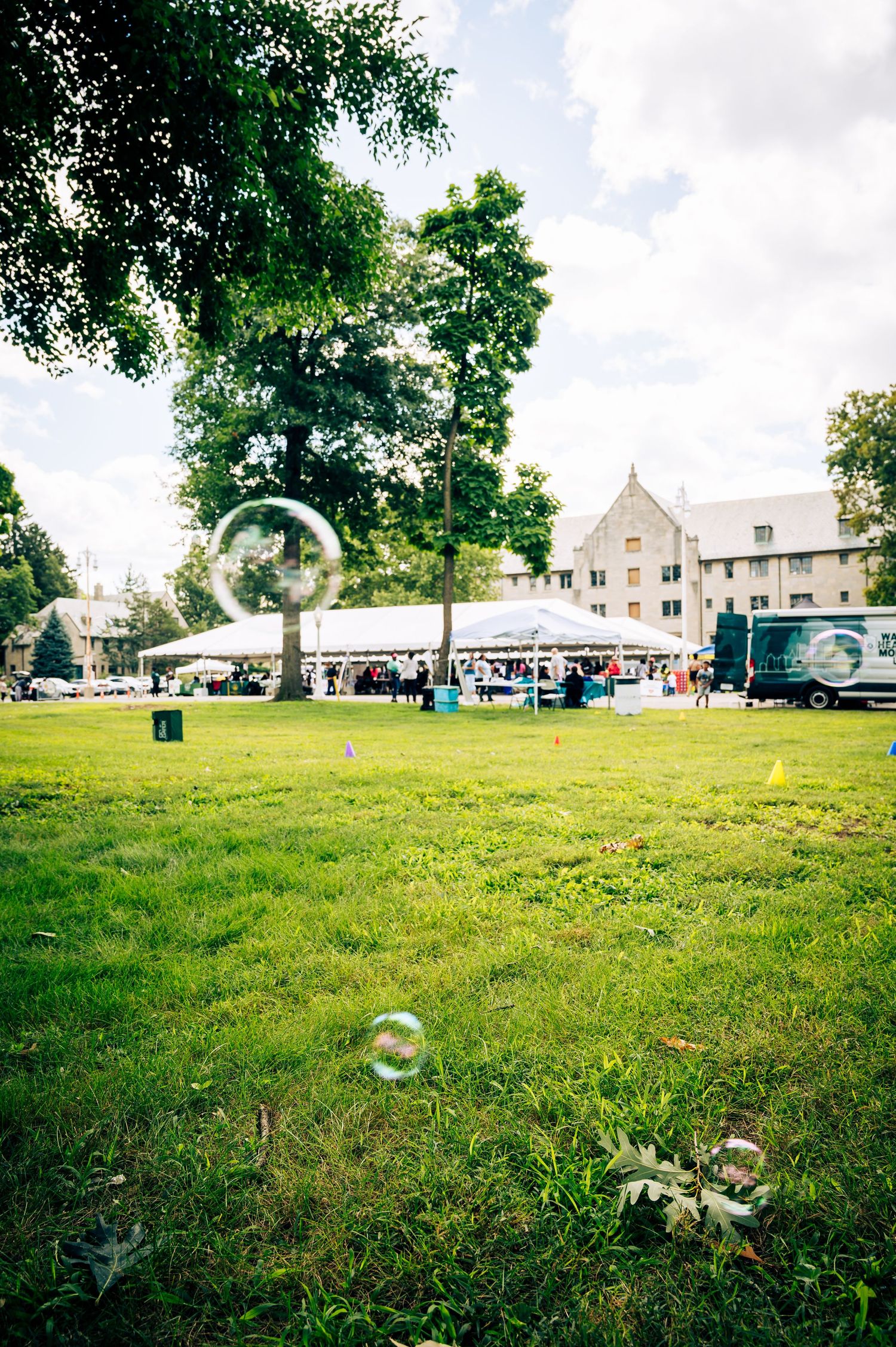 Outdoor event tent on green lawn with large soap bubbles floating in foreground, trees and stone building in background.