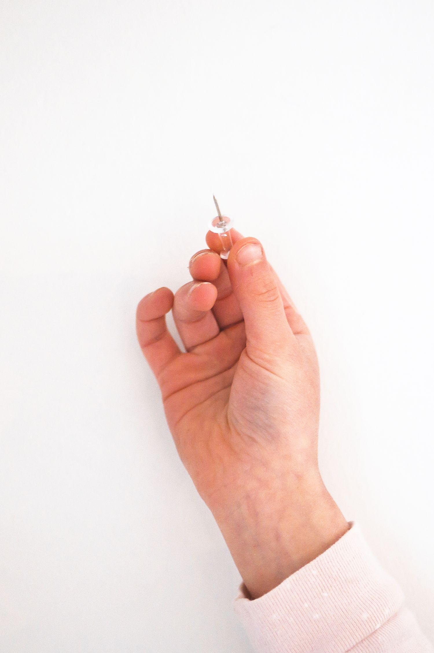 A hand pressing a small pink pushpin into a white wall surface.