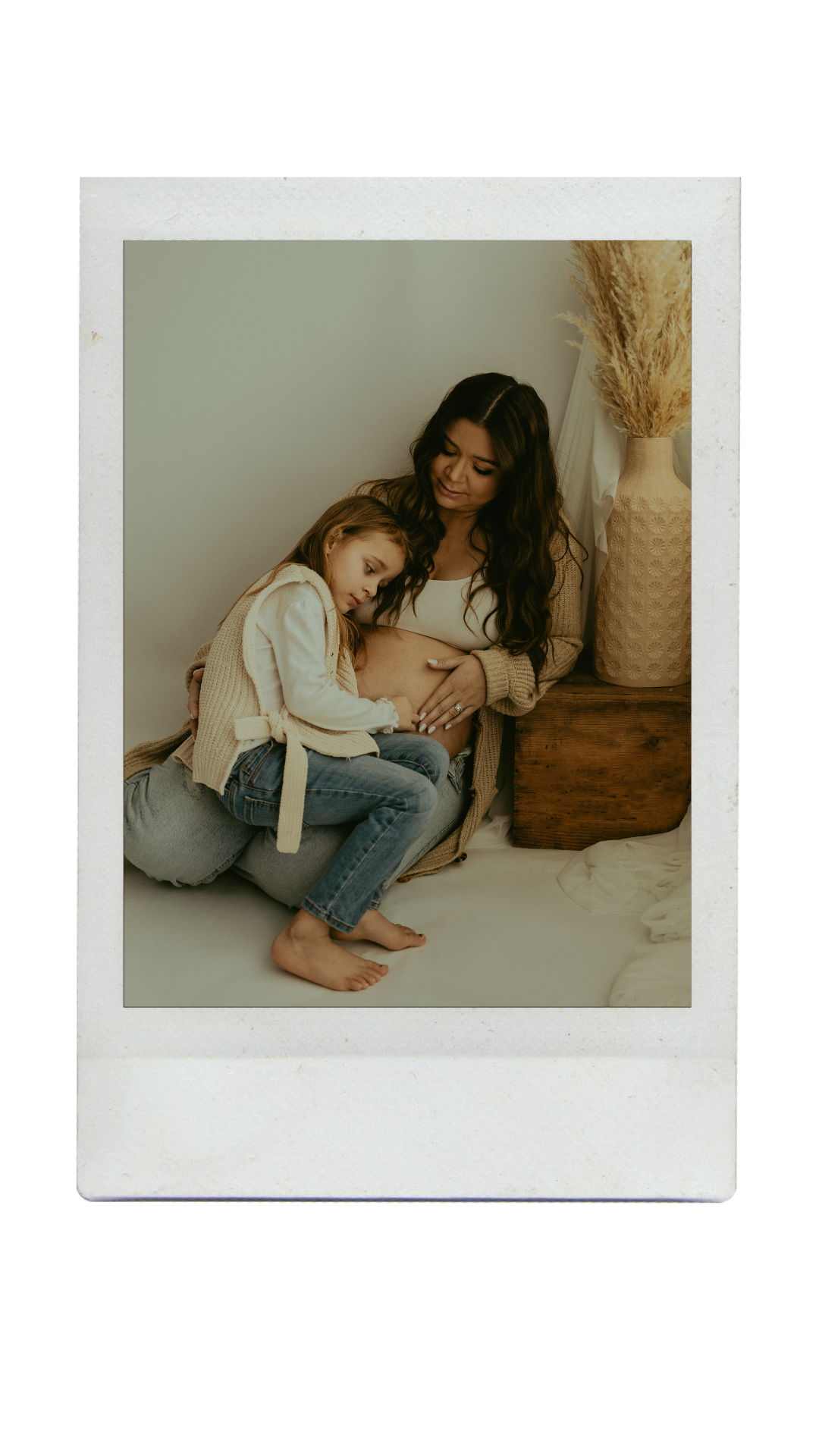 A tender moment captured between mother and child sitting together on the floor in a minimalist white room with pampas grass decor.
