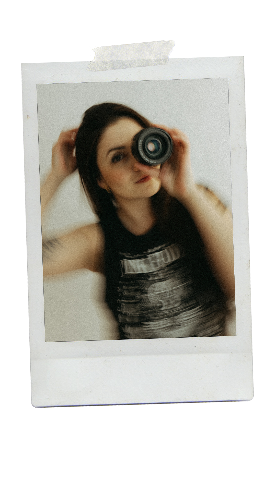 A photographer looks through a camera lens while wearing a dark sleeveless top in a vintage Polaroid-style photo.