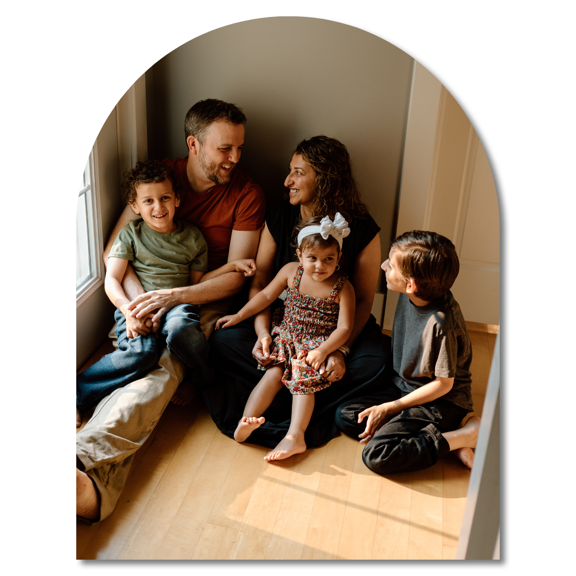A family sitting together in a window nook sharing smiles and laughter in warm natural lighting.