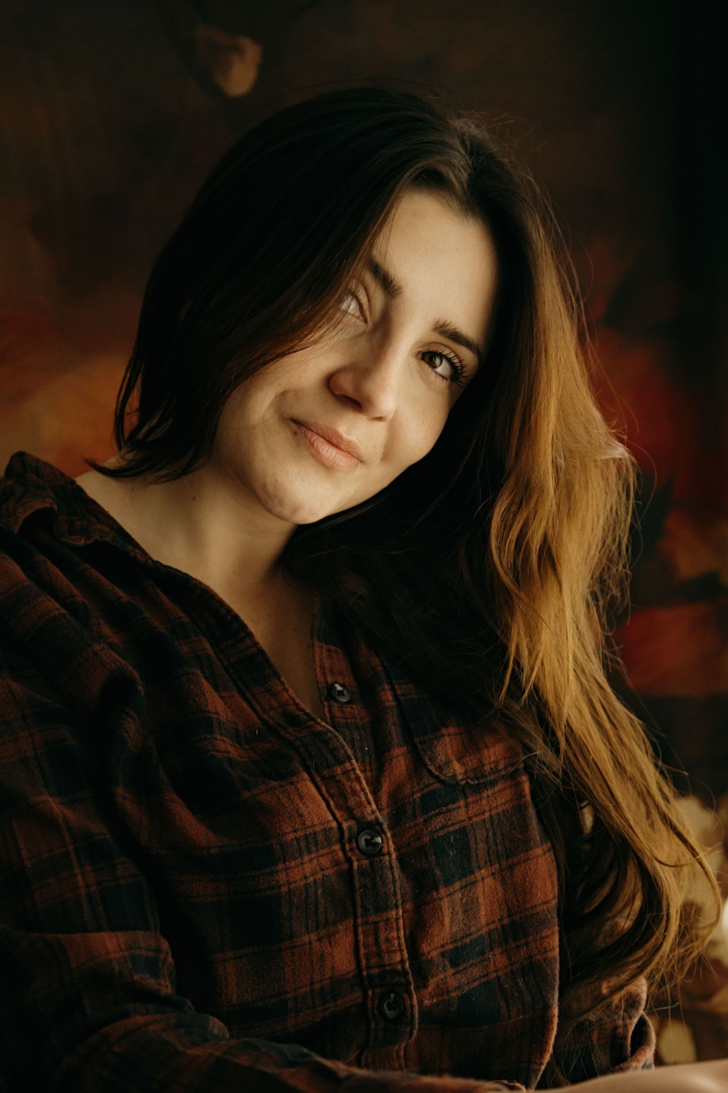 A warm portrait featuring long brown hair and a cozy brown plaid flannel shirt against a dark moody background.