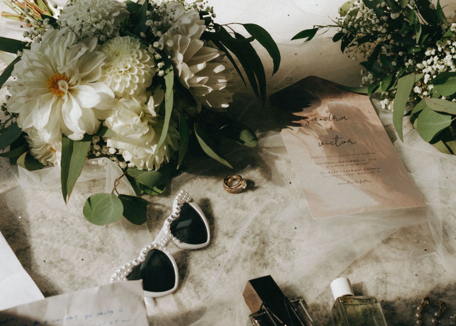 Vintage-style wedding flat lay with white peonies, heart-shaped sunglasses and greenery on beige fabric.