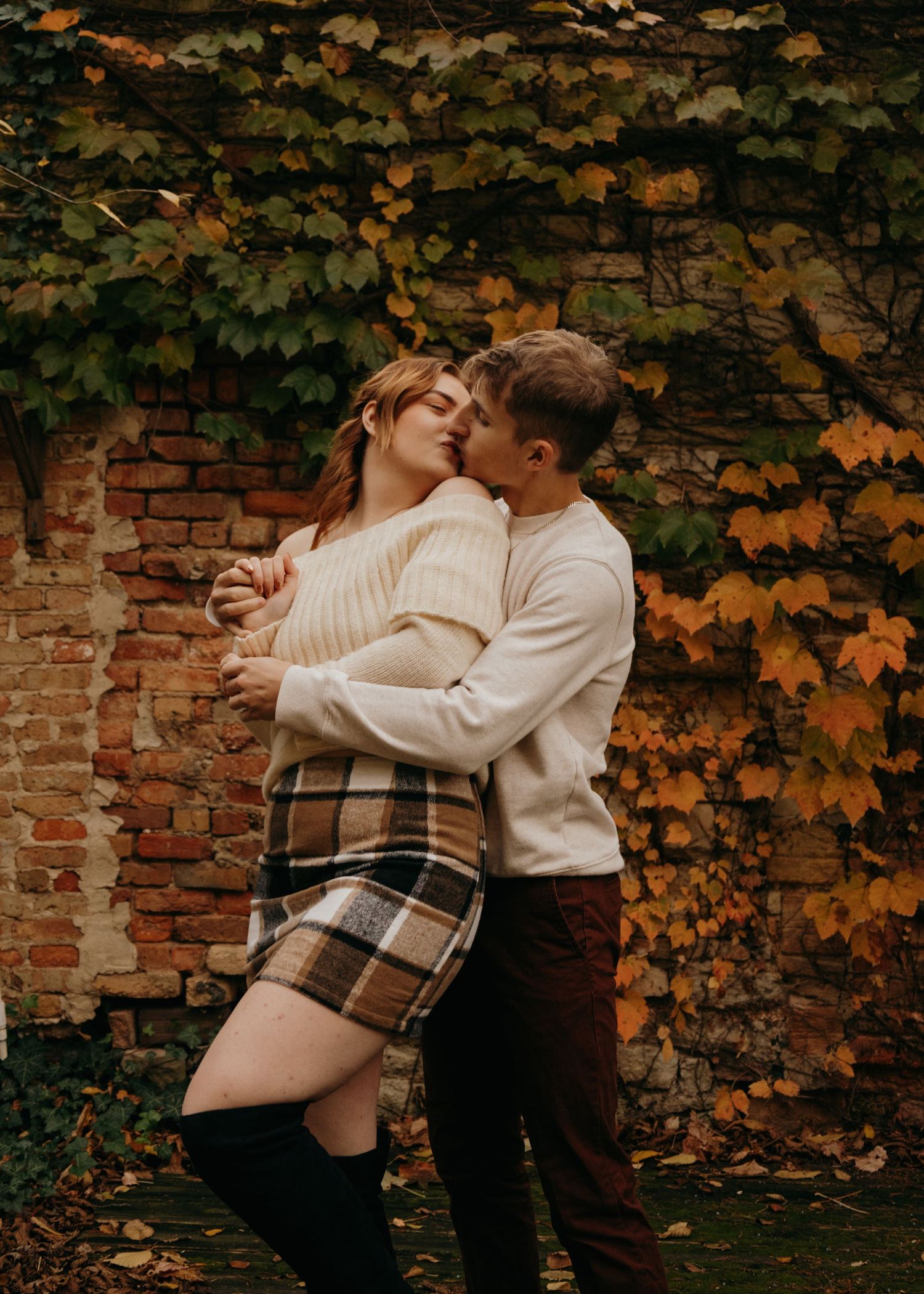 A romantic autumn scene against a brick wall with ivy, featuring warm cream sweaters and a plaid skirt.