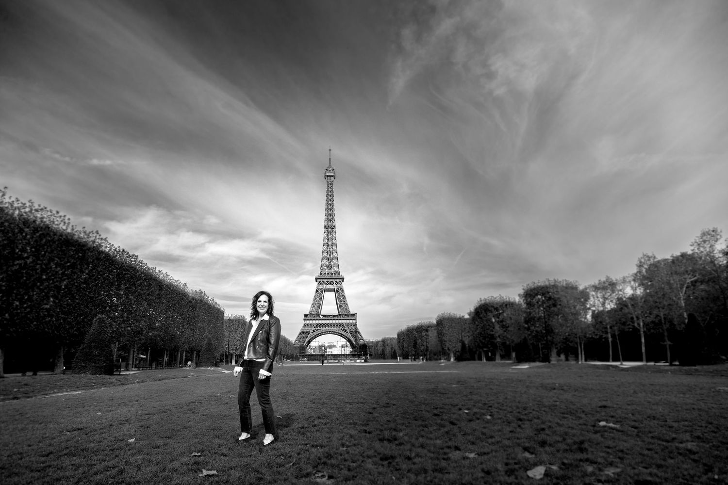 uma mullher de aproximadamente 60 anos está em pé no Campo de Marte em Paris, ao fundo, centralizada na foto, se vê a Torre Eiffel