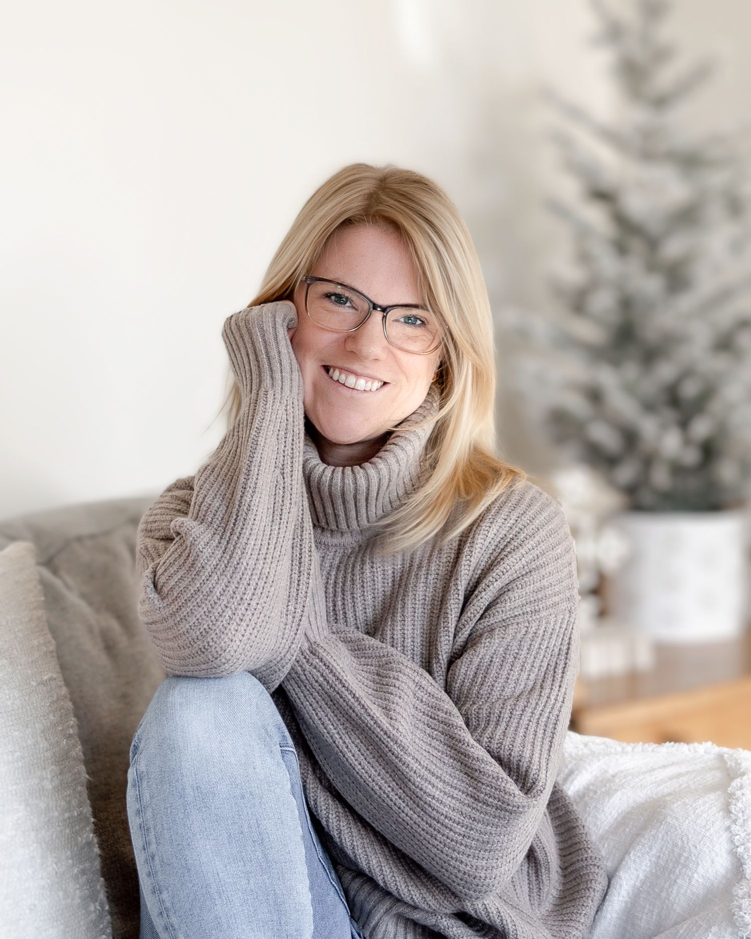 Person in cozy gray turtleneck sweater relaxes on white couch with snowy winter scene visible through window.