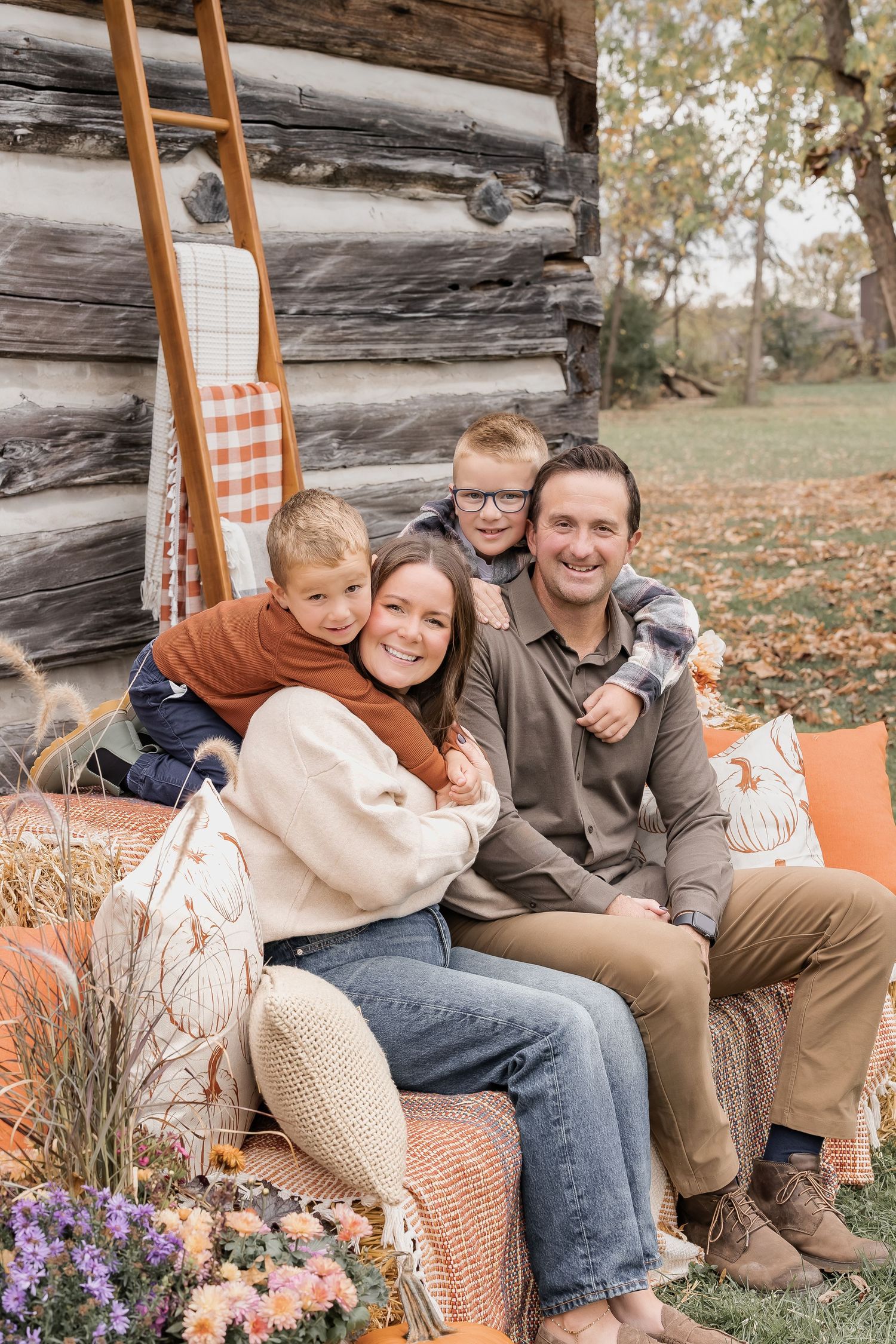A family sits together on rustic wooden steps surrounded by fall decor and autumn leaves in a cozy outdoor setting.