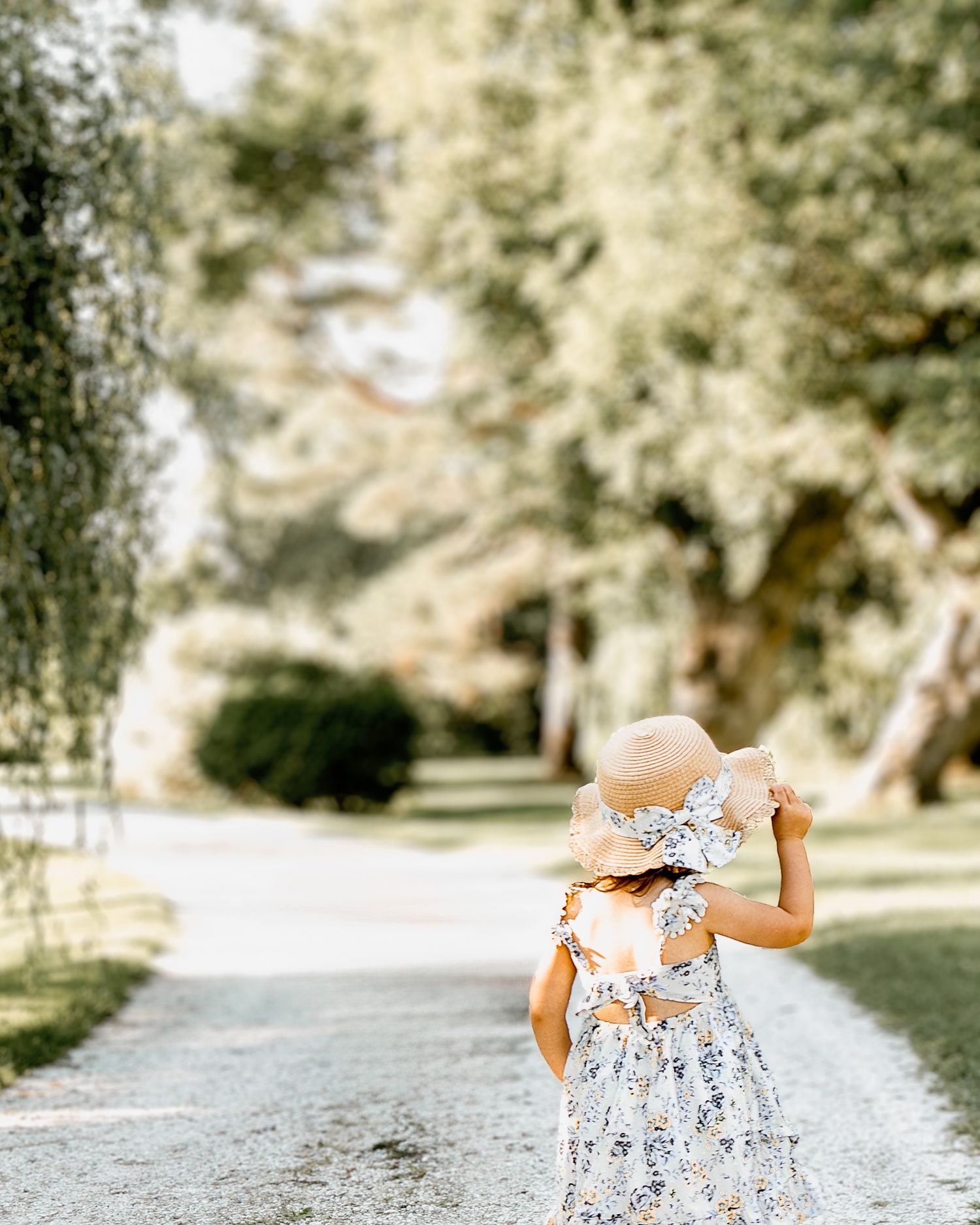 A person in a floral dress walks down a tree-lined path in a park on a sunny day.