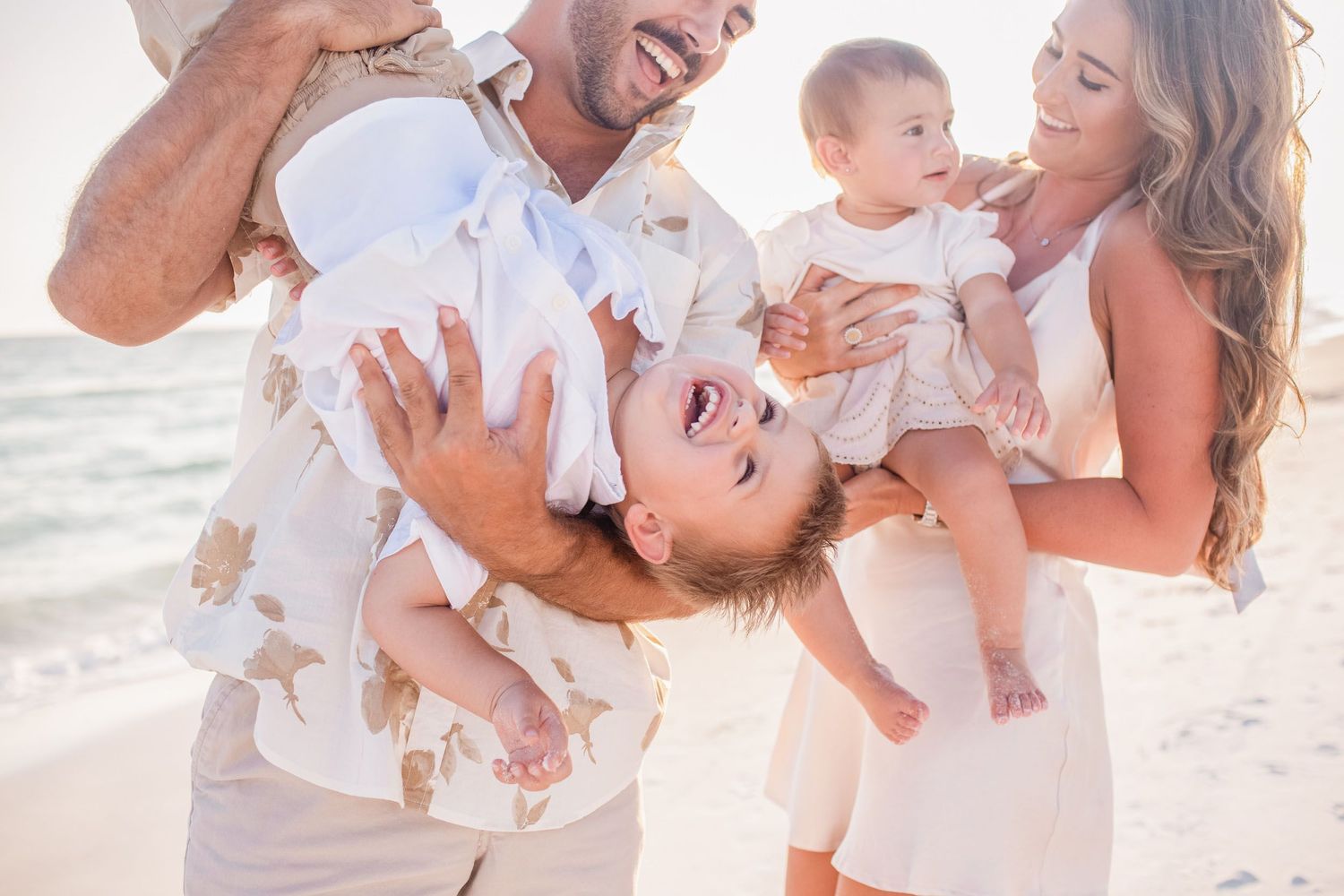 A joyful family wearing white outfits plays together on a sunny beach during a photo session.