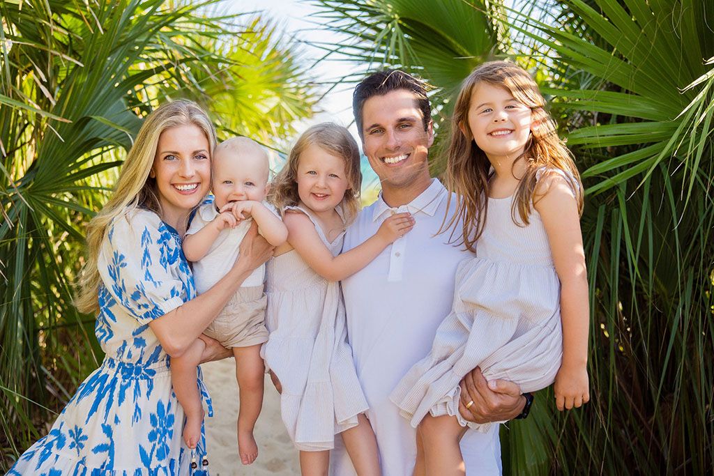 A family wearing white and blue outfits poses together outdoors among tropical palm fronds in bright sunlight.