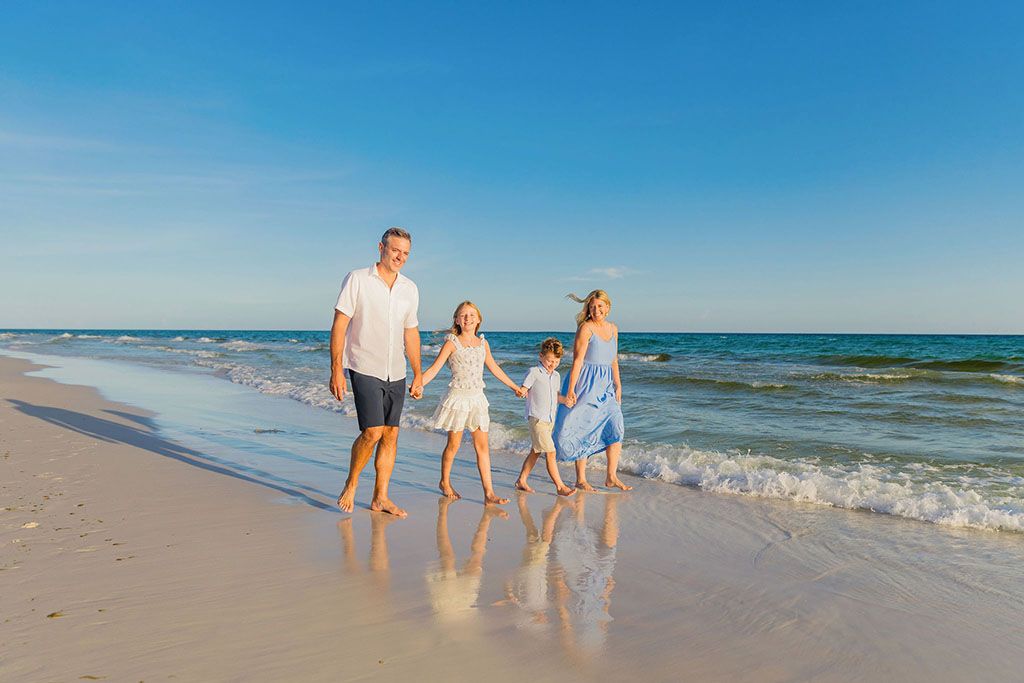 Family walking together and holding hands along a sunny beach shoreline with ocean waves and blue sky in the background.