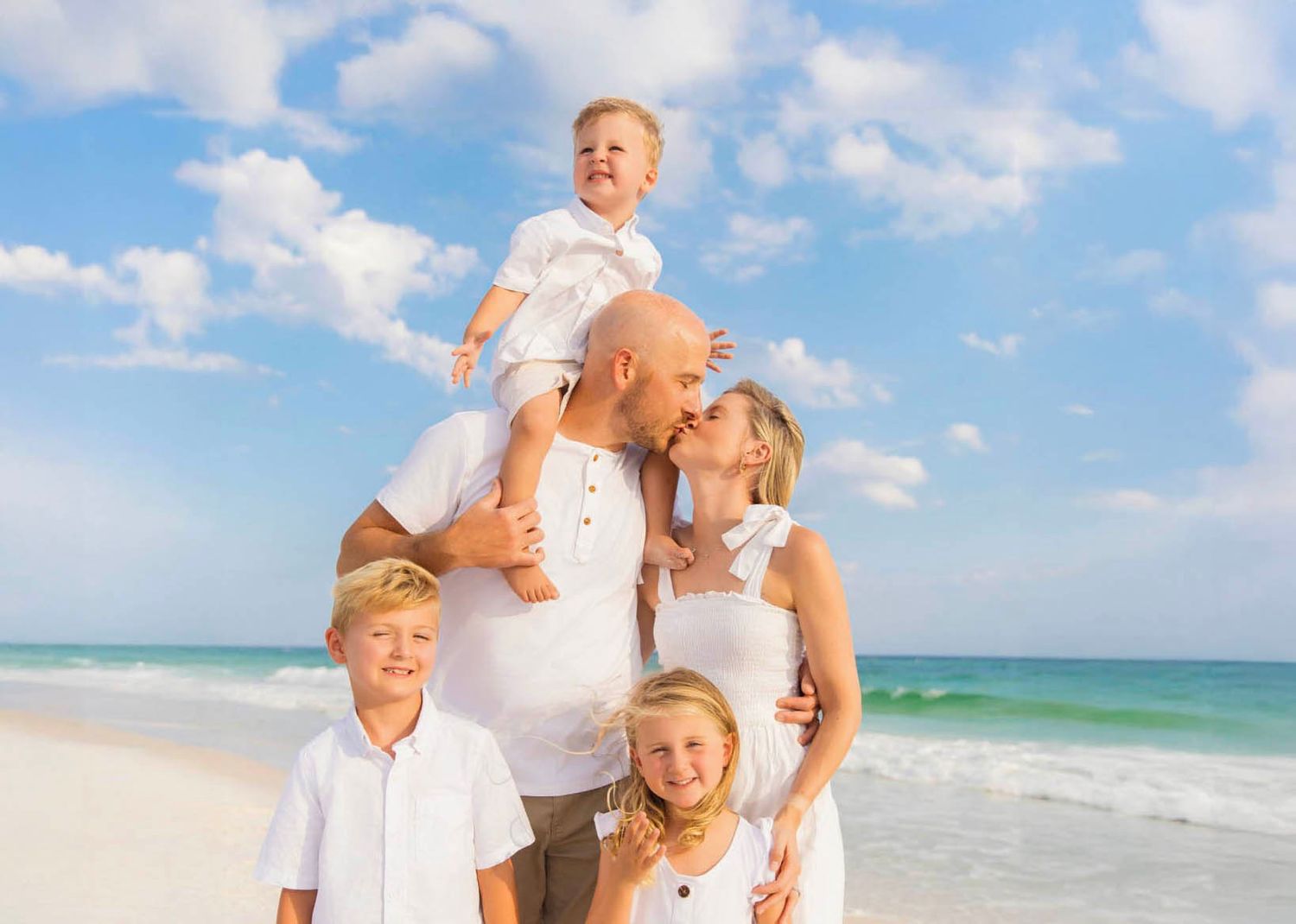 A family wearing white outfits poses together on a sunny beach with turquoise ocean waves in the background.