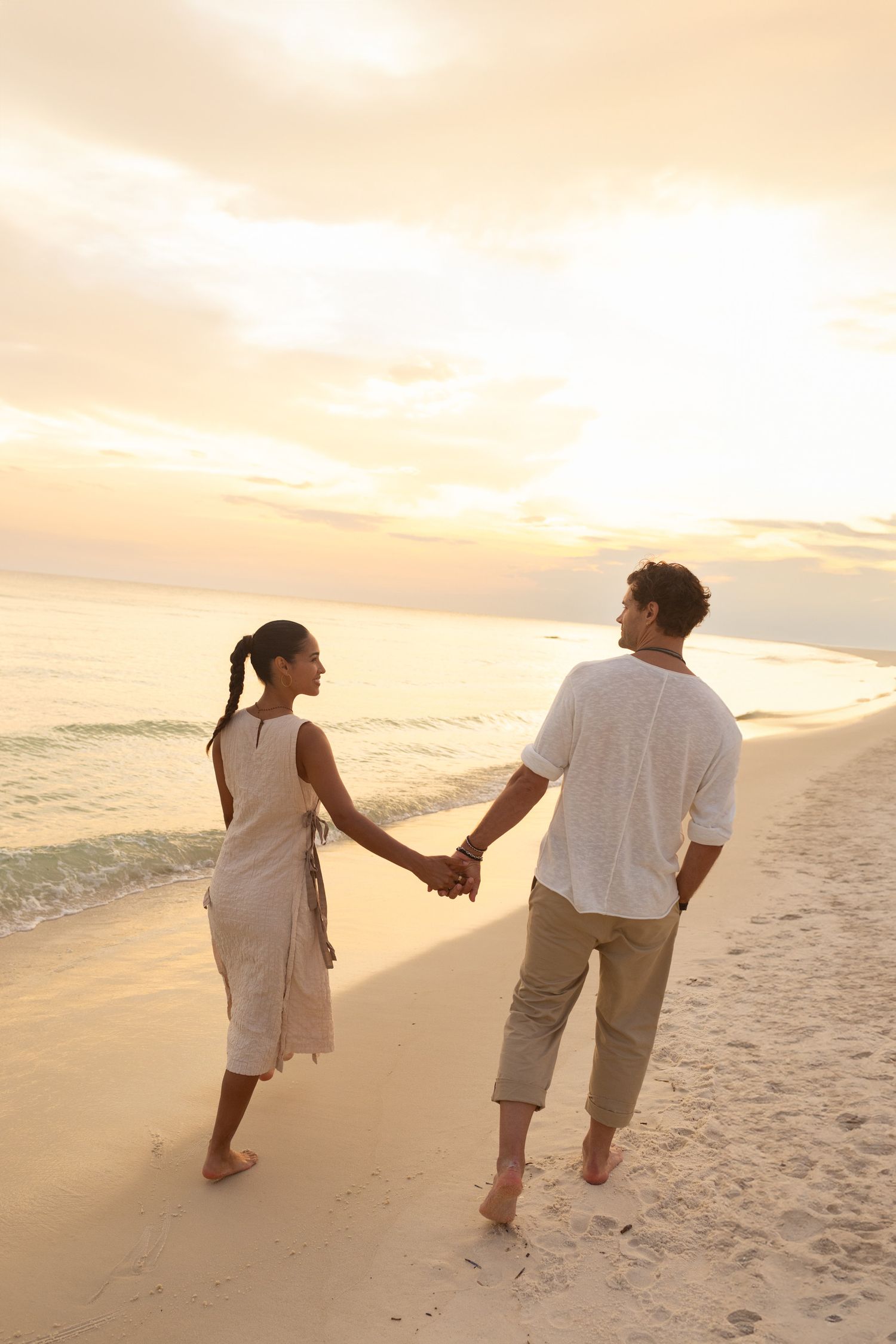 A couple walks hand in hand along a sandy beach at sunset with warm golden light illuminating their path.