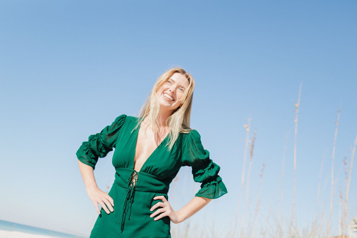 Woman in emerald green dress poses against blue sky and beach grass in a summer outdoor photoshoot.