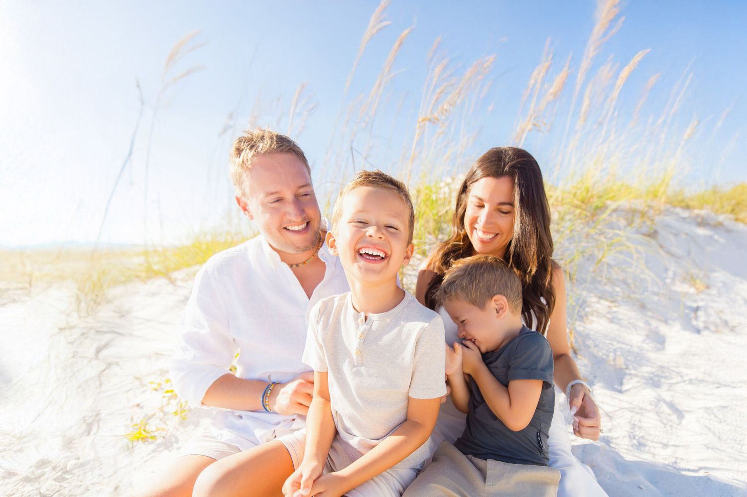 A family sits together on a sunny beach with tall grass and sand dunes in the background.