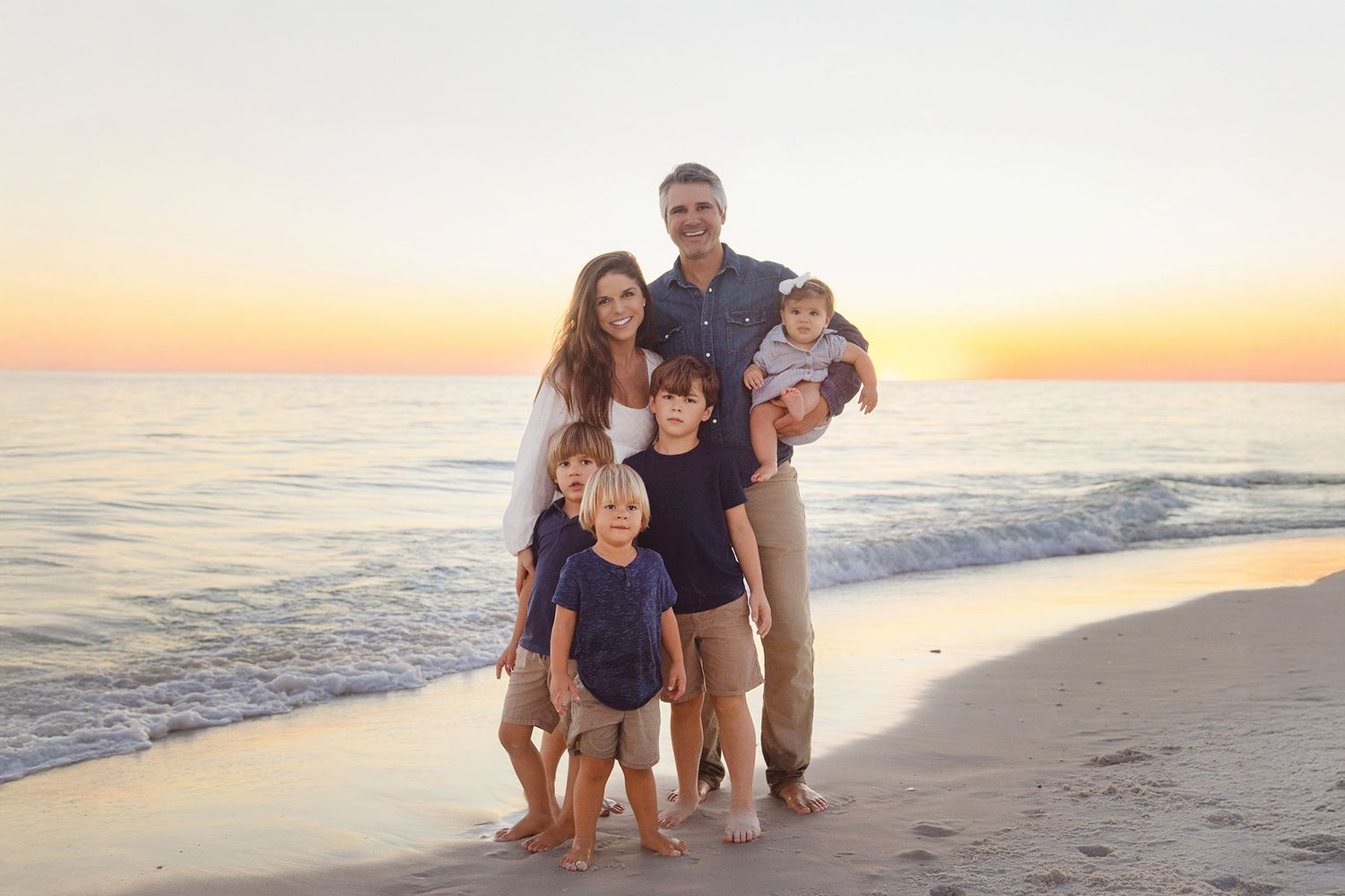 A family stands together on a beach at sunset with the ocean and golden sky in the background.