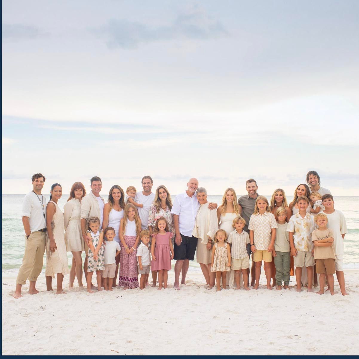 Large family group wearing coordinated neutral outfits poses together on a sunny beach at sunset.