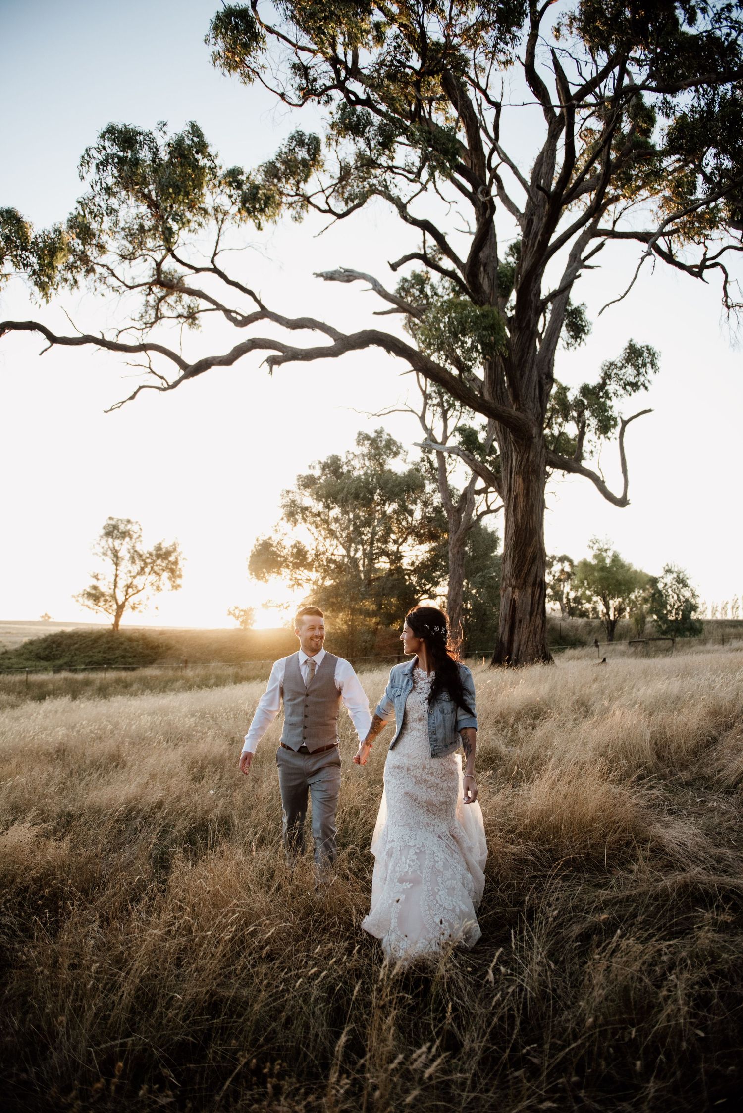 Newlyweds walk through tall grass beneath towering eucalyptus tree at sunset.
