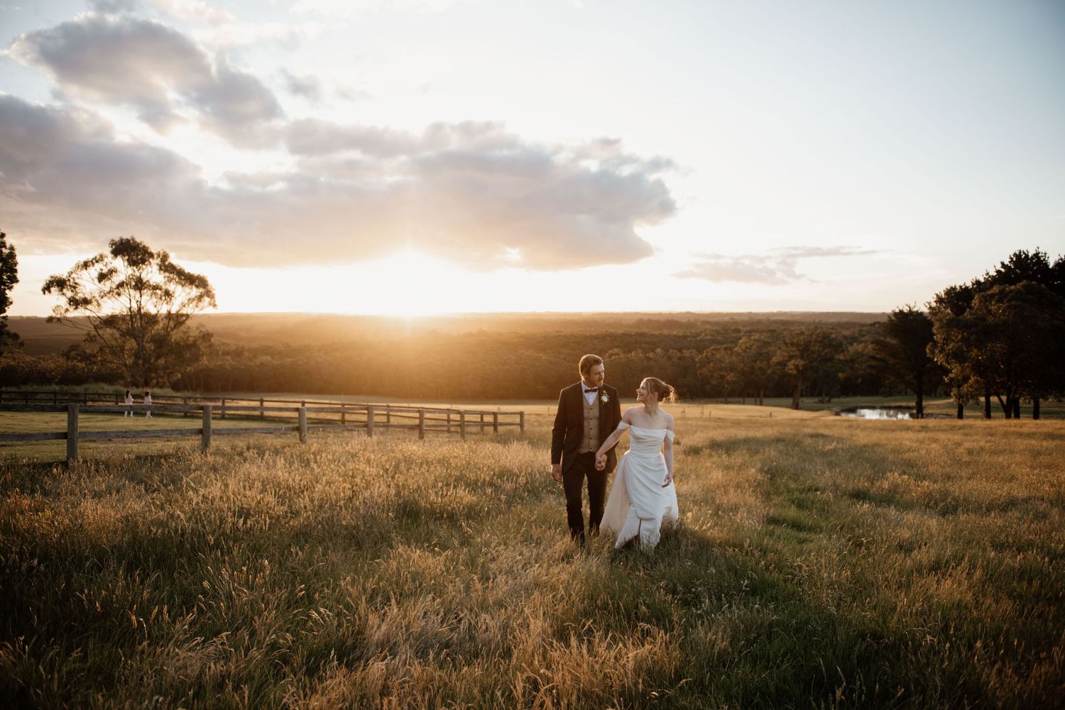 Newlyweds walk hand in hand through a golden field at sunset.