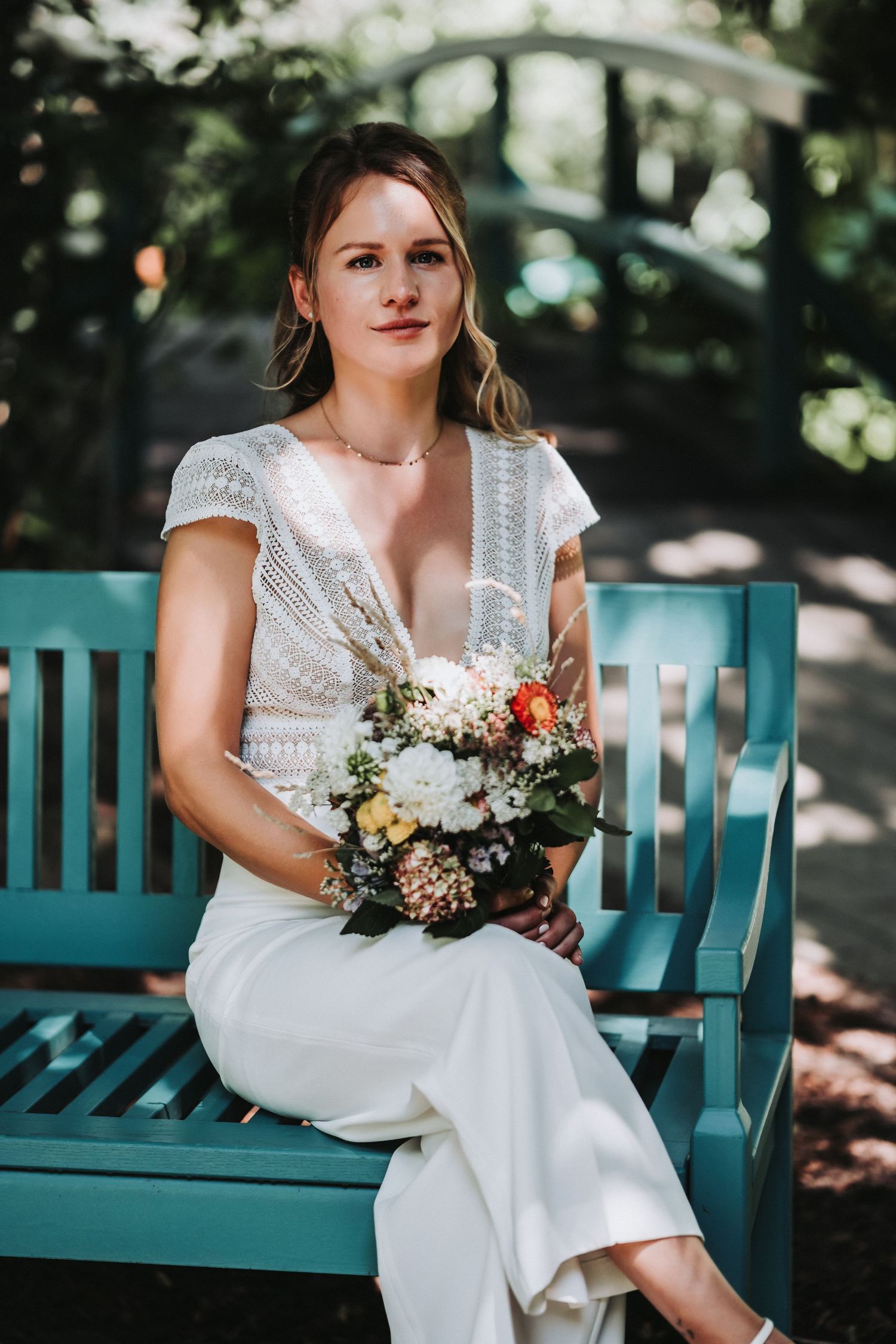 A bride in a white dress sits on a teal-colored garden bench holding a colorful bouquet in a sunlit outdoor setting.