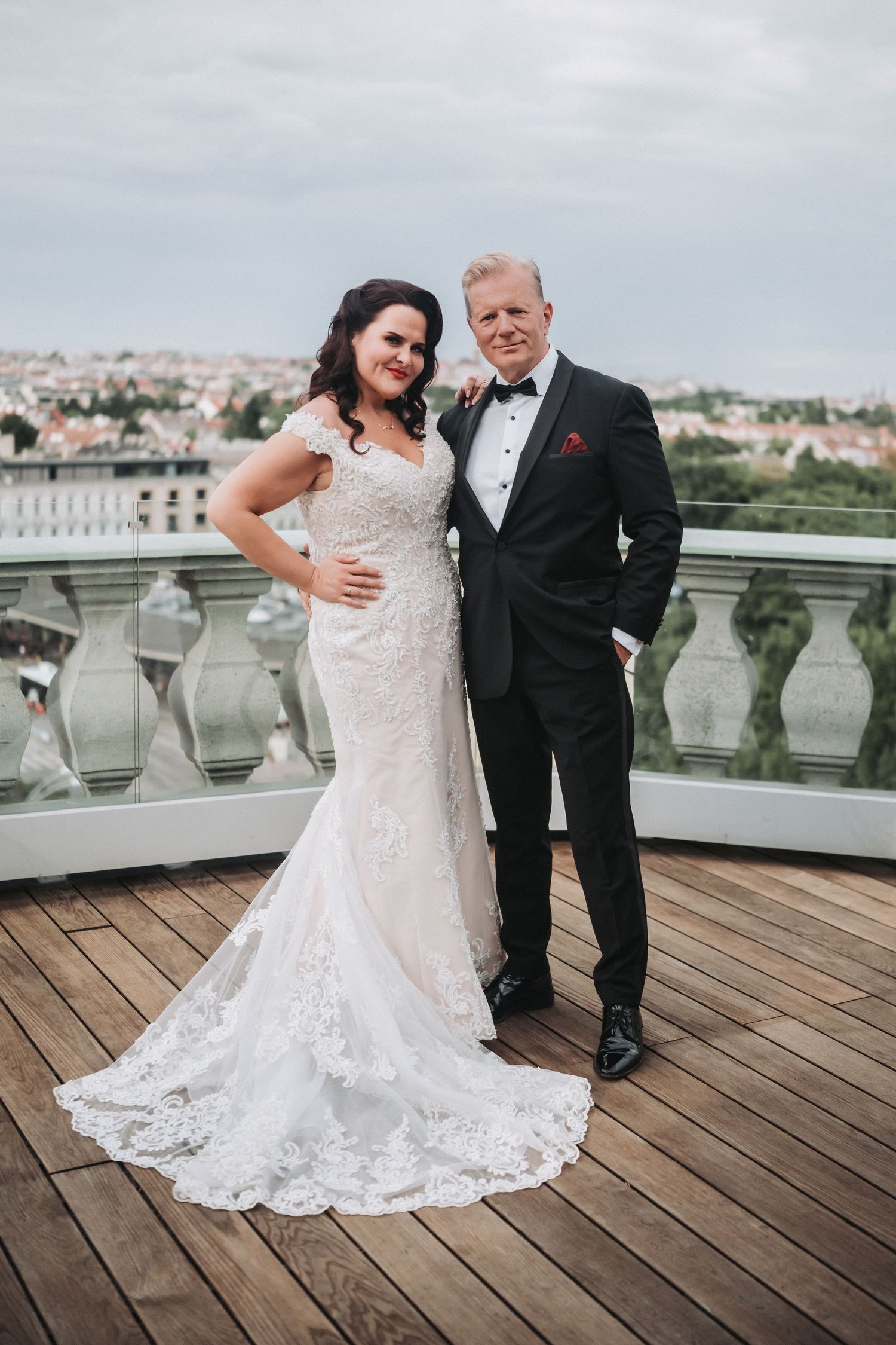 Wedding couple poses on a wooden deck with ornate stone balustrade and city views in the background.