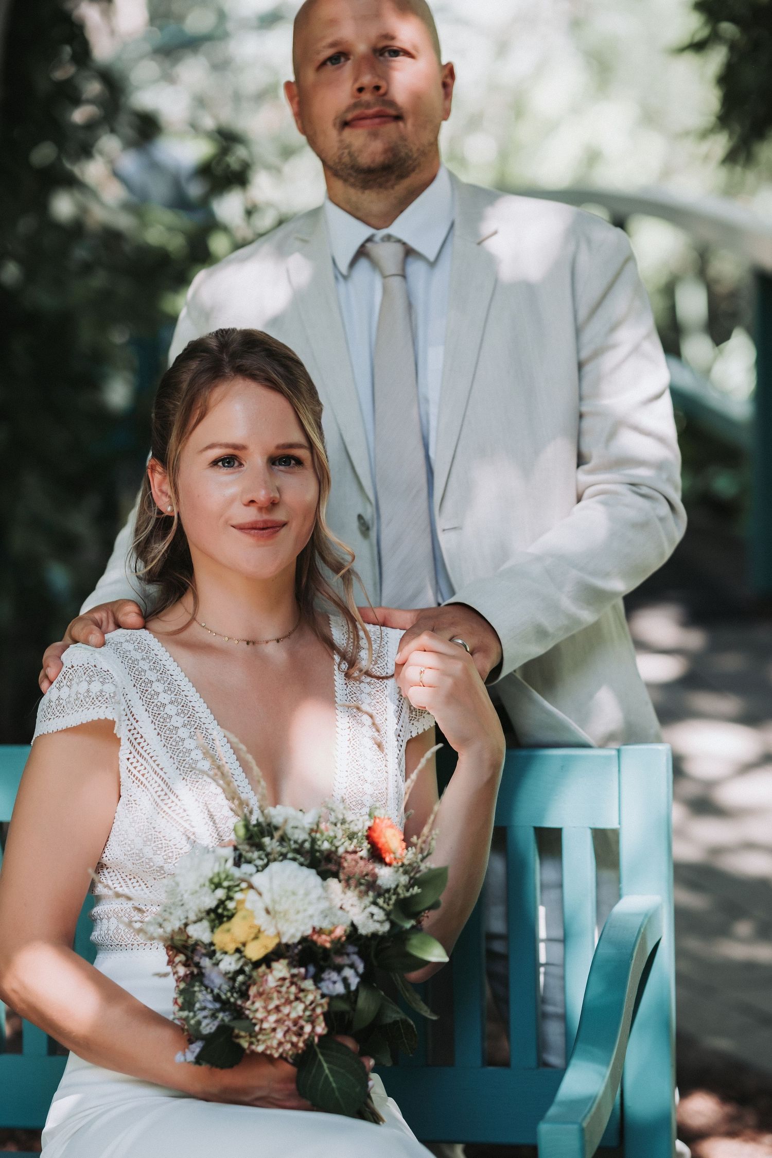 Couple poses on turquoise bench during outdoor wedding photoshoot with bride holding delicate floral bouquet.
